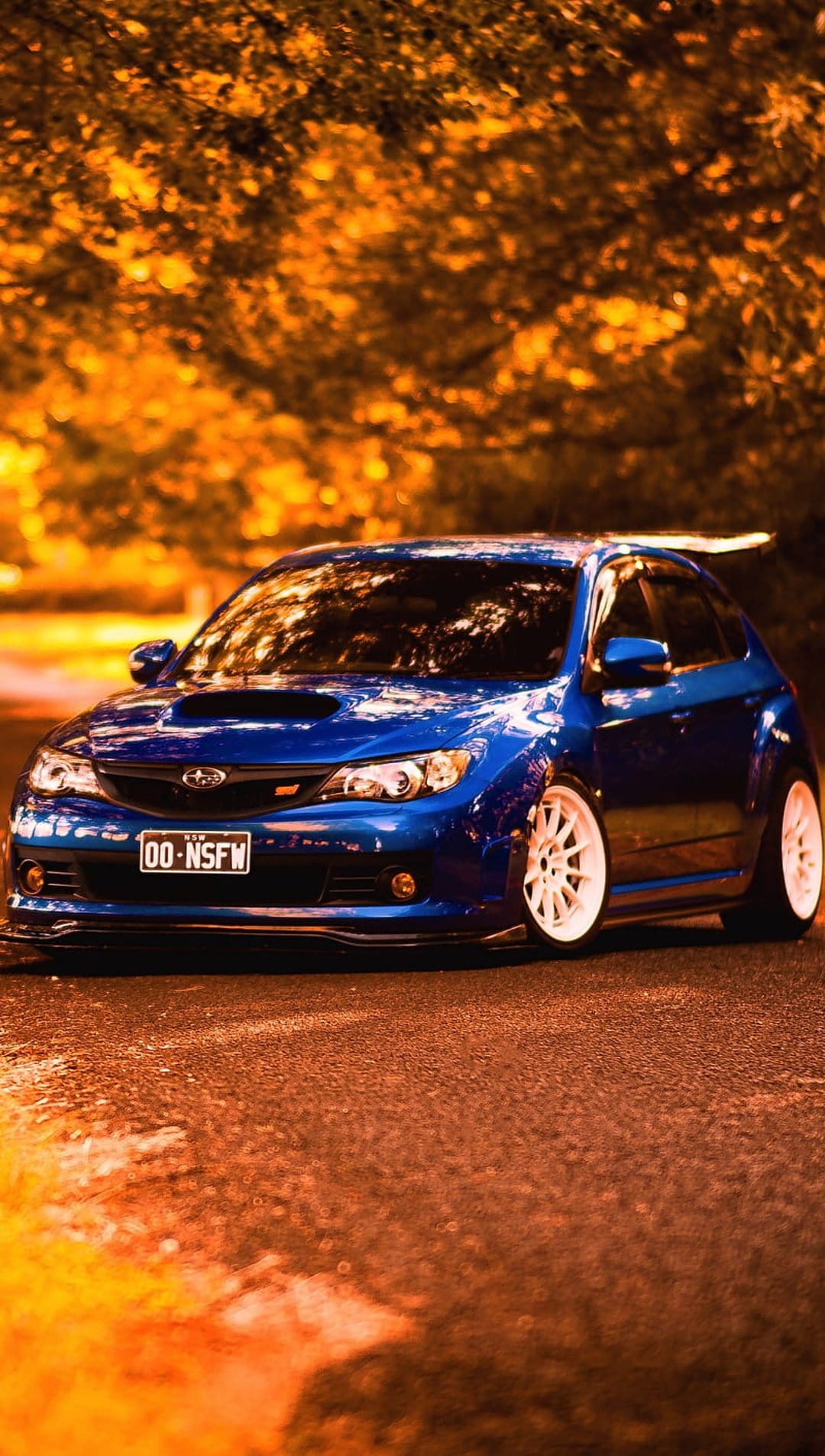 Blue Subaru WRX STI hatchback with white wheels on wet pavement with golden autumn trees in the background.