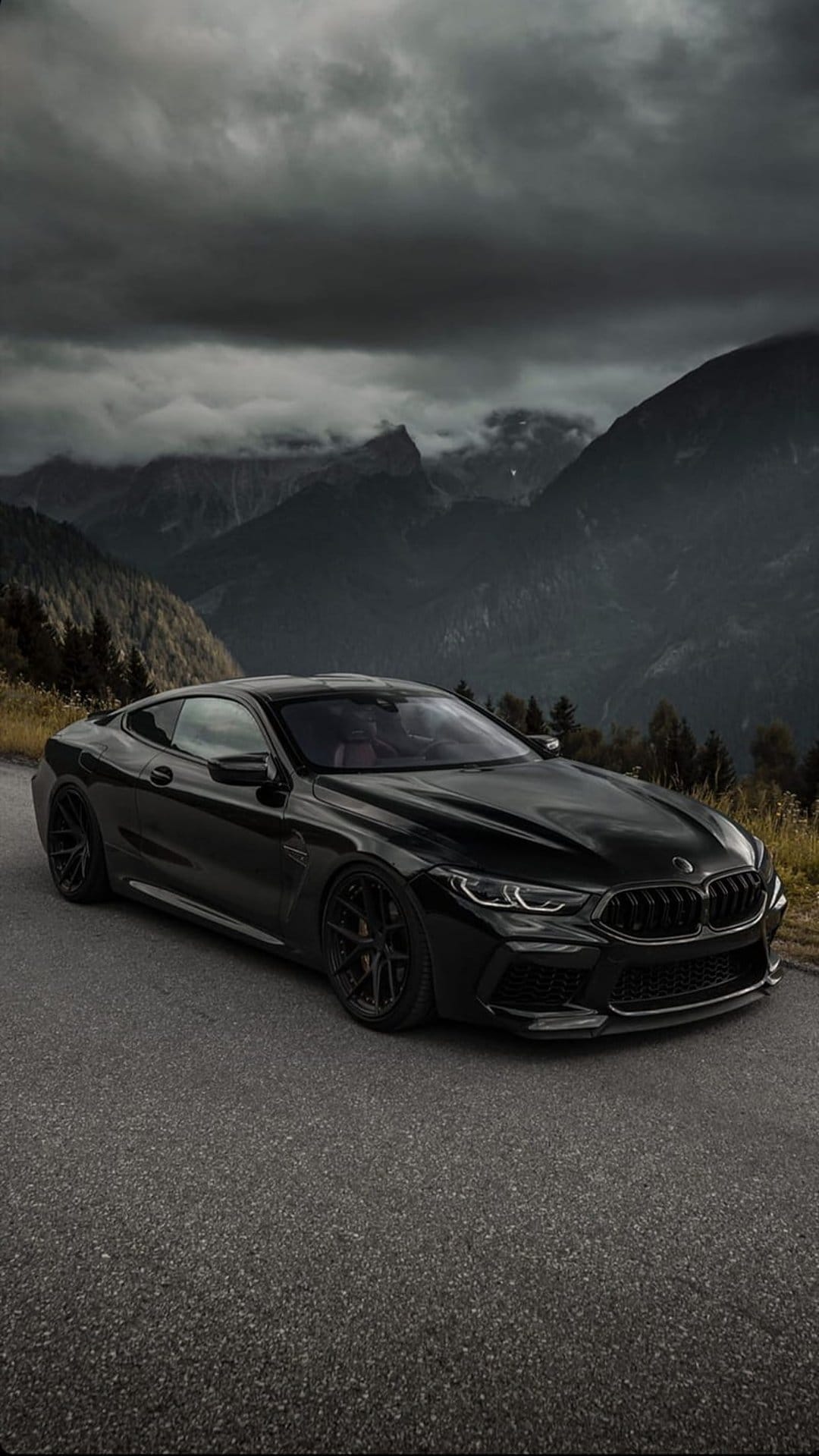 Black BMW M8 coupe parked on a mountain road with dark storm clouds and jagged peaks in the background.