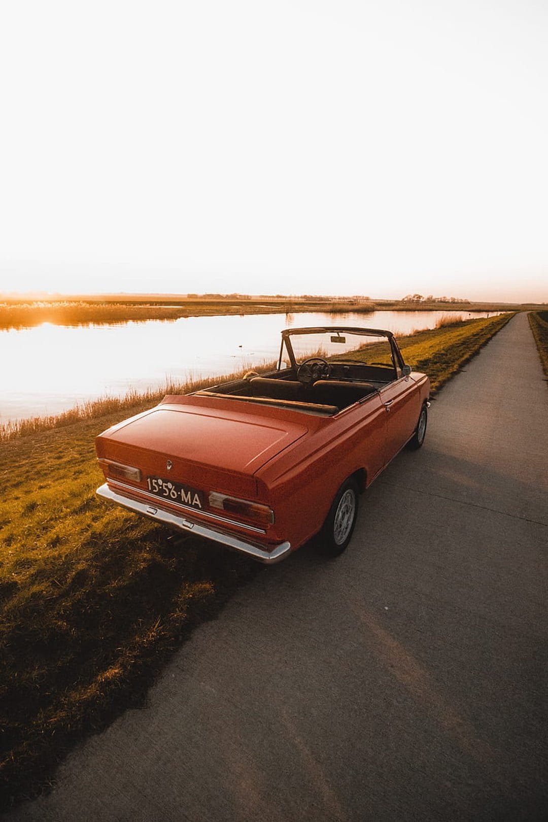 Red vintage convertible parked on a paved path beside a river during a vibrant golden hour sunset at dusk.