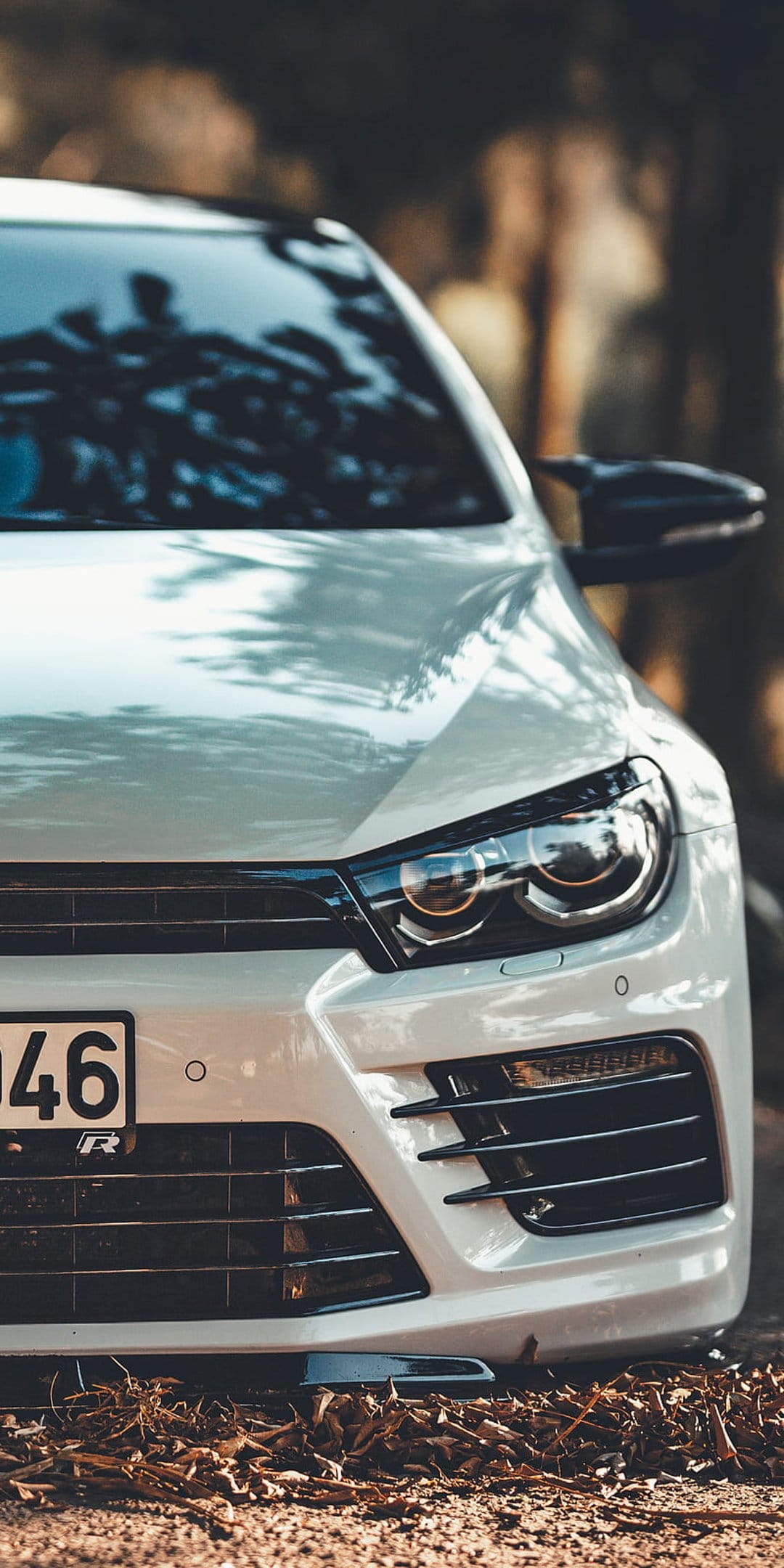White Volkswagen Scirocco front bumper and headlights resting on dry orange autumn leaves in a blurred forest park.