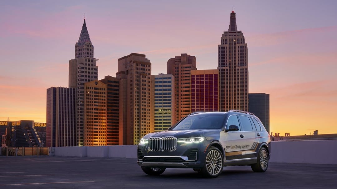 Dark BMW X7 SUV with headlights on a rooftop against a New York-style city skyline at purple dusk.