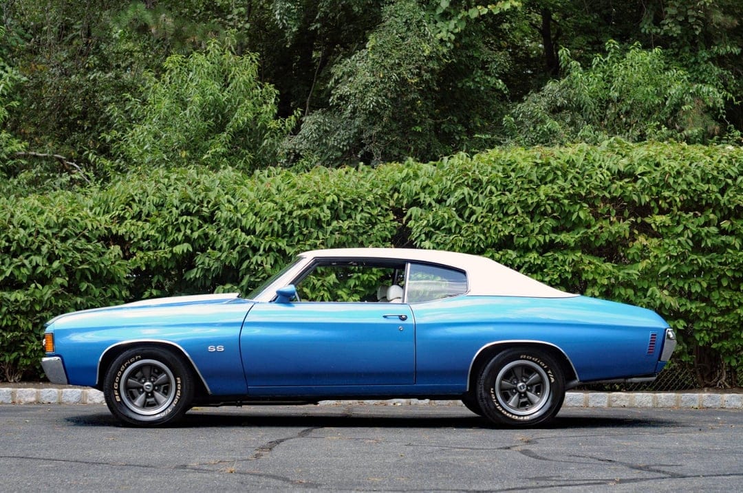 Side profile of a blue 1972 Chevrolet Chevelle SS with a white vinyl top parked on asphalt against green trees.