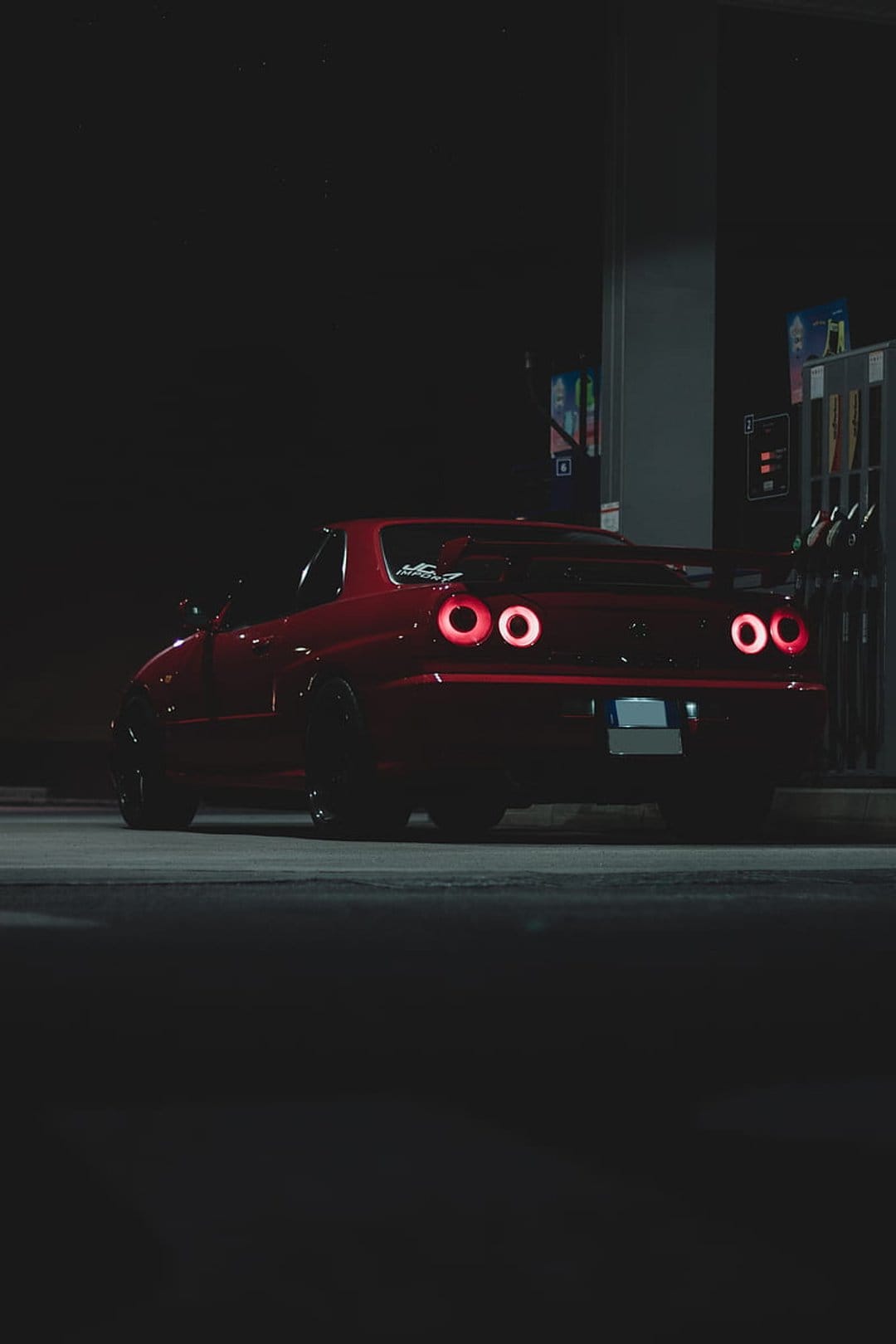 Scarlet supercar with glowing taillights parked at a dark gas station under bright canopy lights at night.