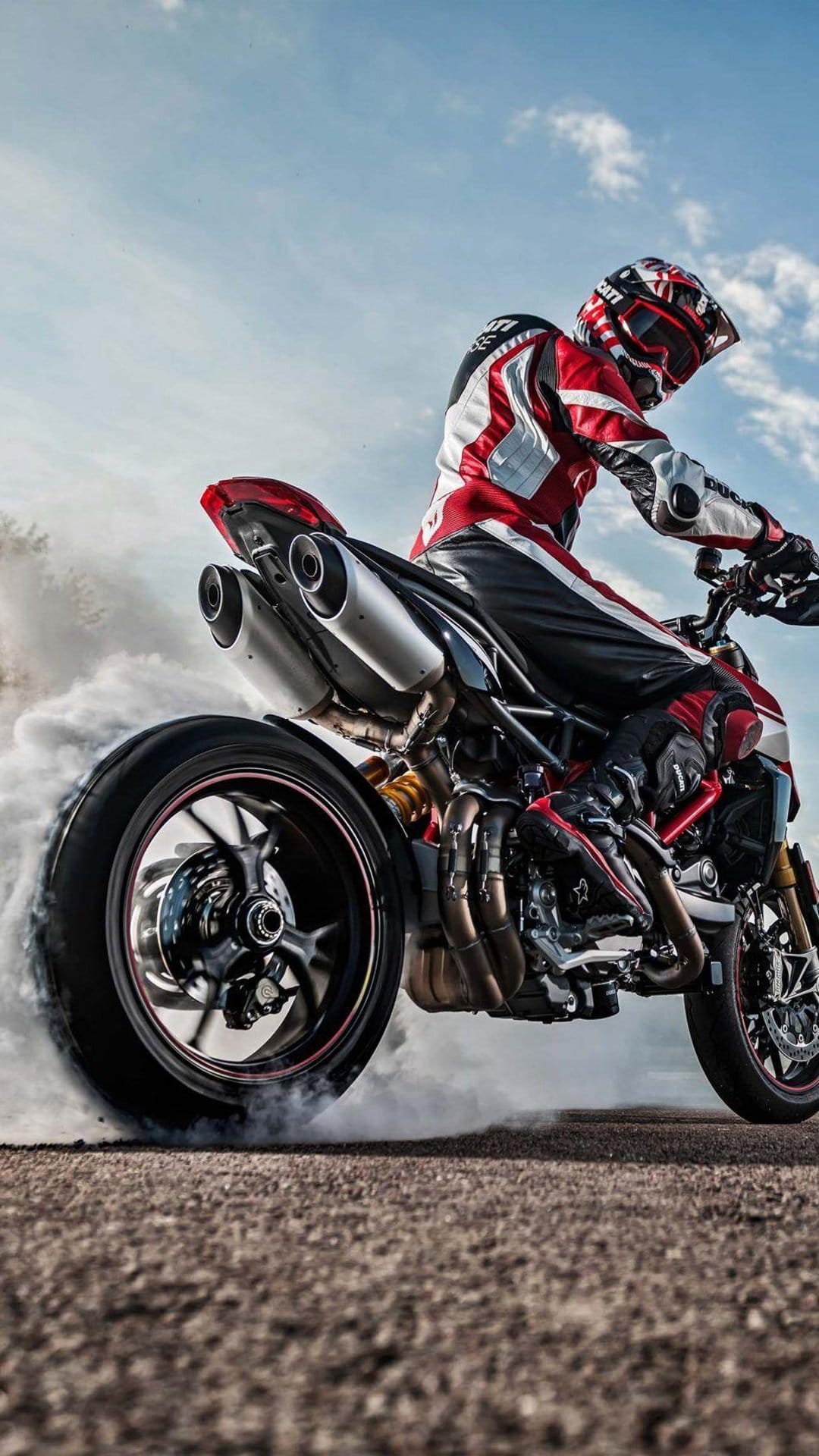Motorcyclist in racing gear performs a burnout with thick white smoke on a street against a bright blue sky.