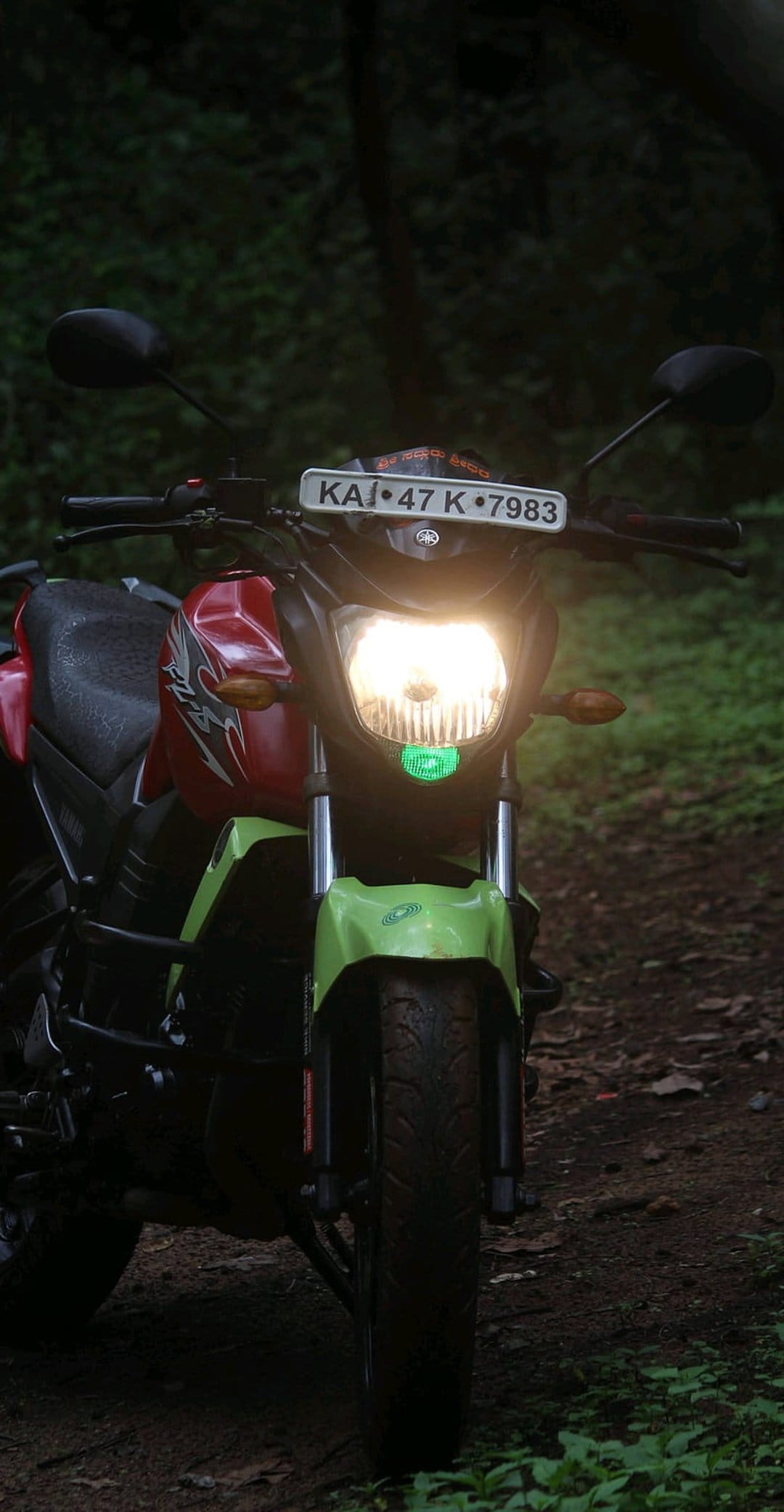 Yamaha motorcycle with glowing headlights parked on a dirt trail surrounded by dark green forest foliage.