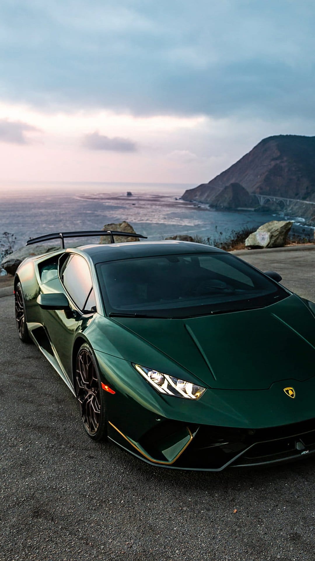 Green Lamborghini parked on a coastal road during a pastel sunset with mountains and a bridge in the distance.
