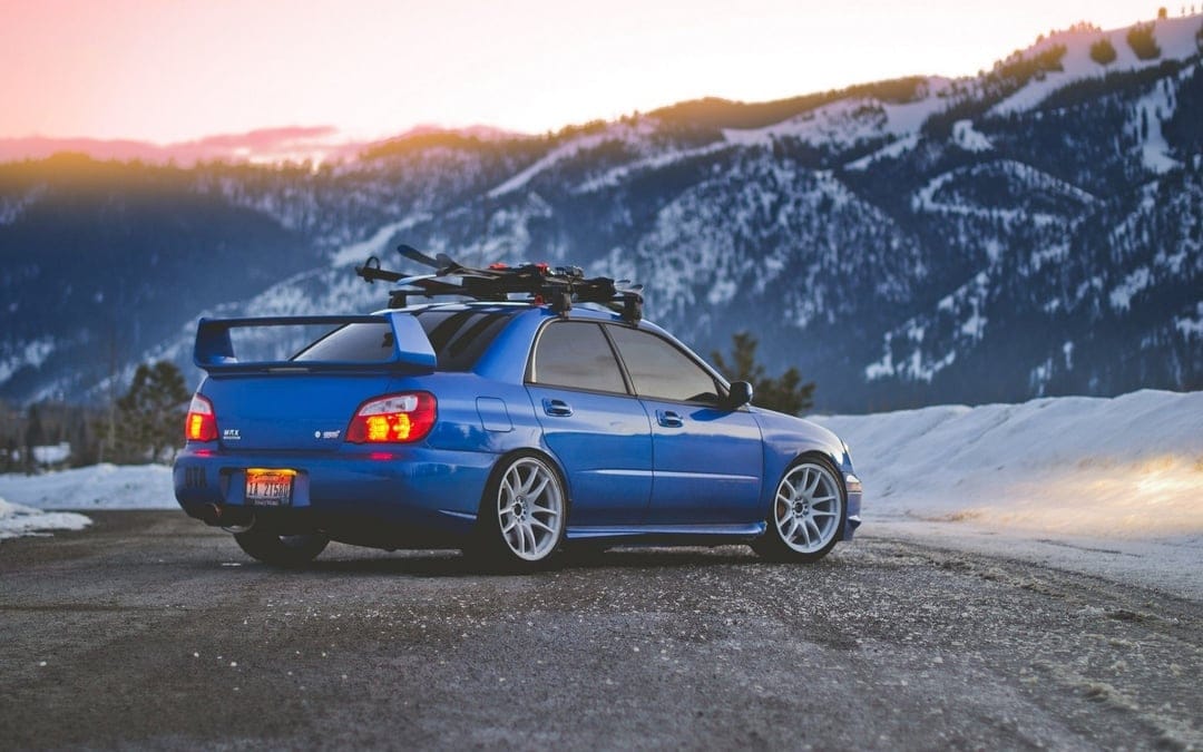Blue Subaru WRX STi with skis on roof parked on a mountain road at sunset against snow-capped peaks.