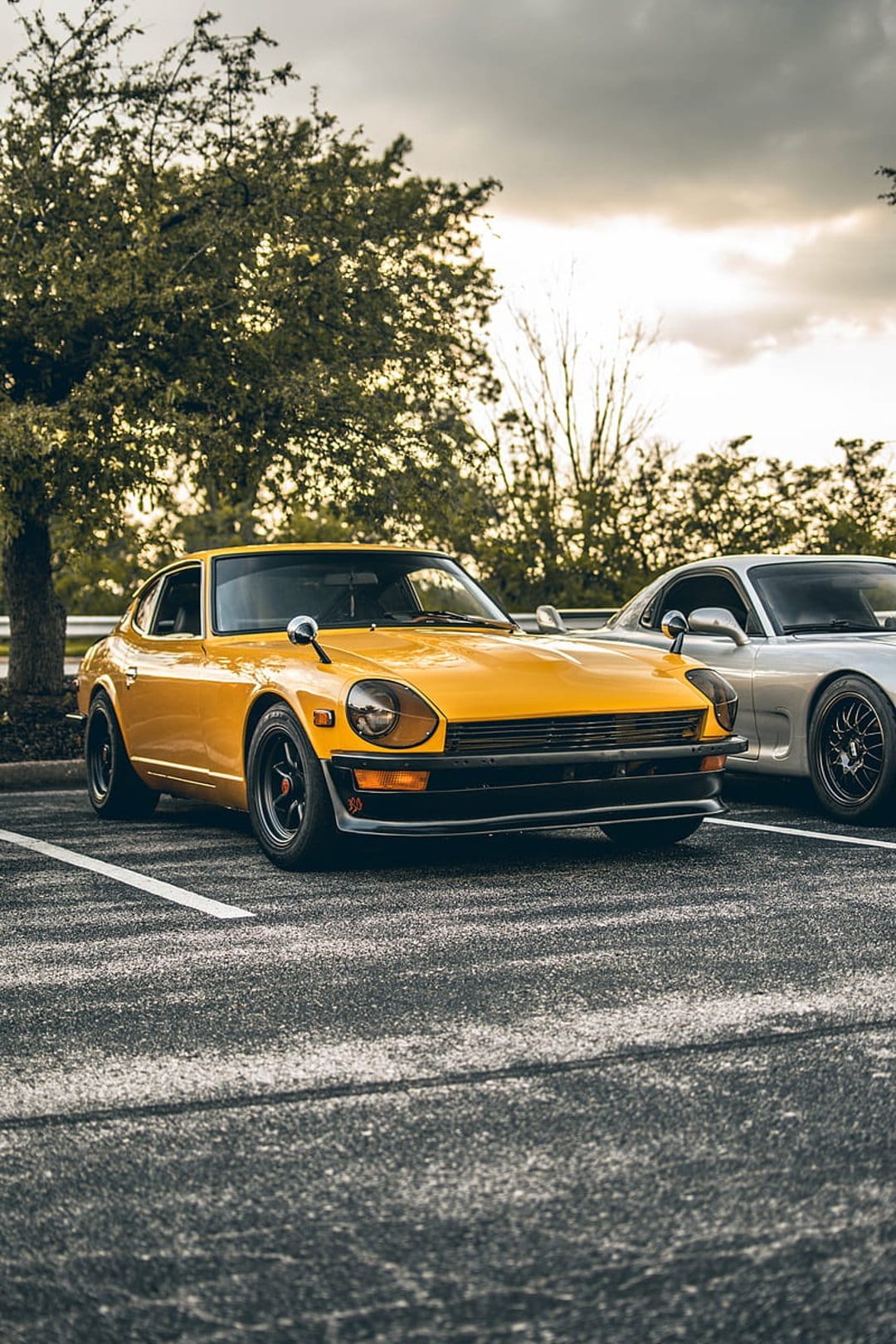 Yellow Datsun 240Z parked next to a silver Mazda RX-7 in a lot with a background of trees under overcast skies.