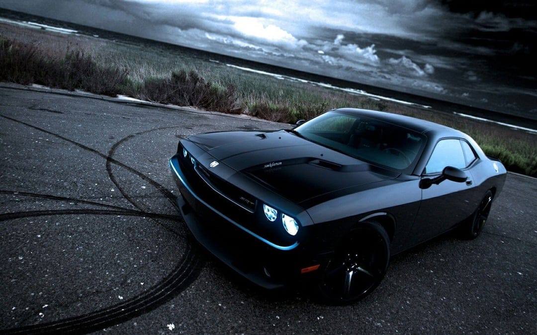 Black Dodge Challenger SRT with glowing headlights on a coastal road under a stormy sky with burnout marks.