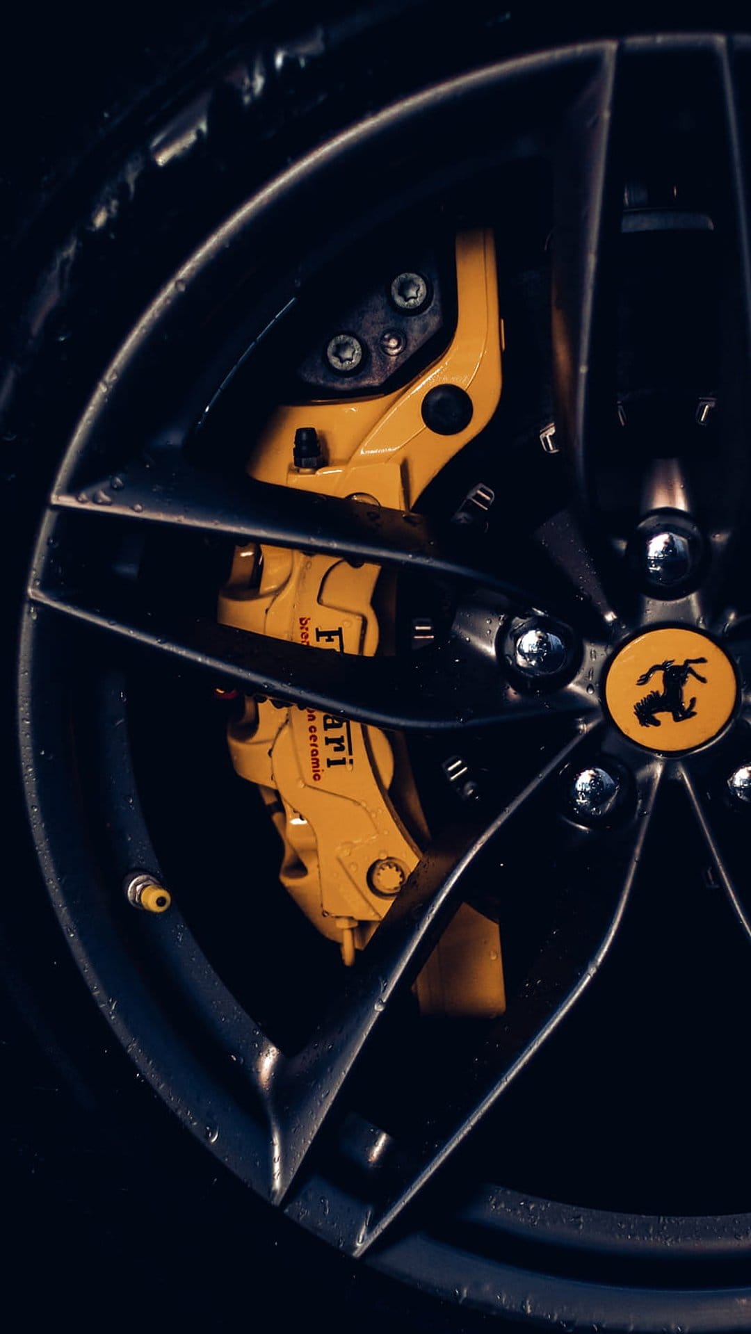 Black Ferrari wheel rim featuring a yellow brake caliper and prancing horse logo with realistic water droplets.