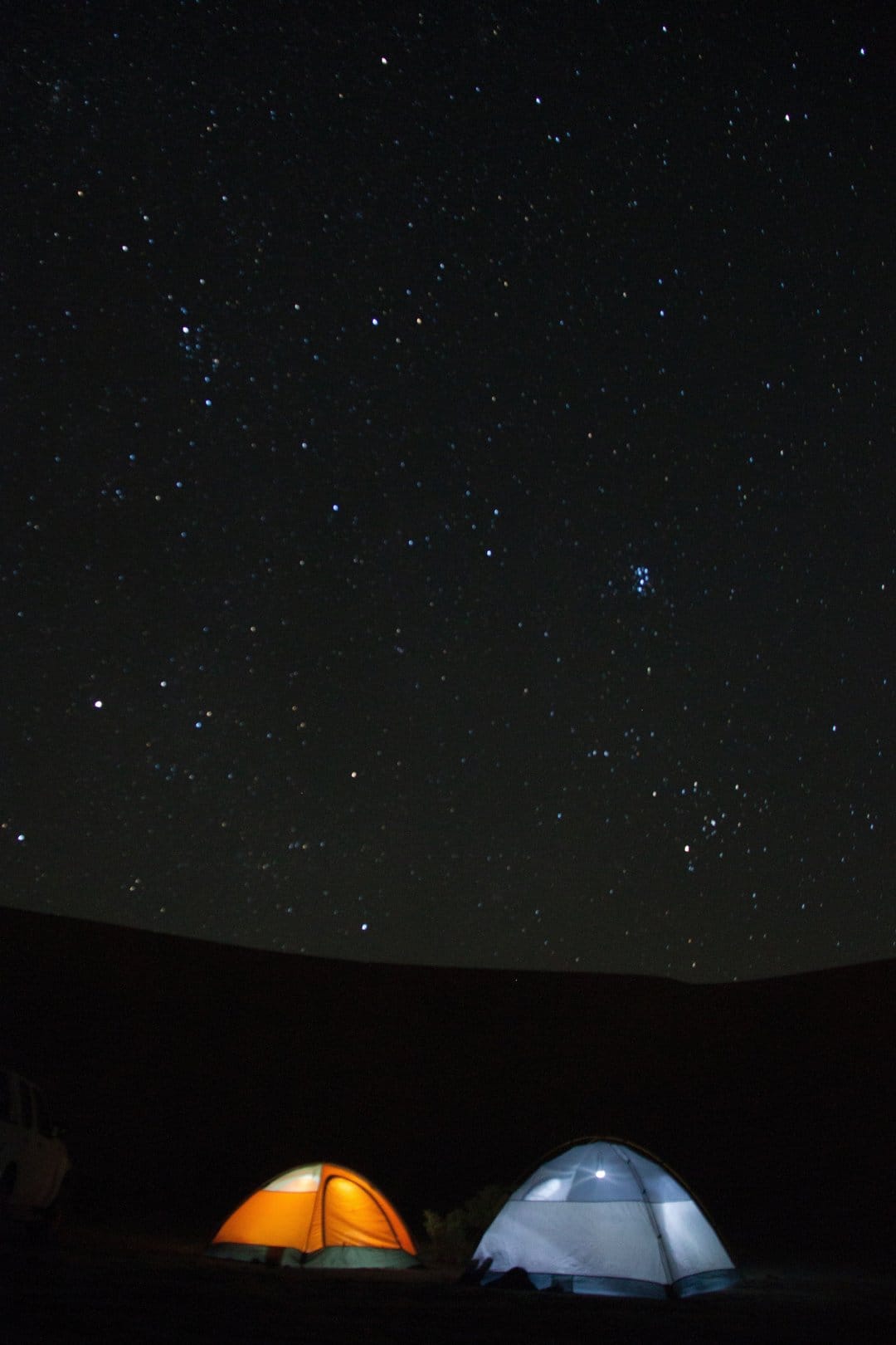 Two glowing tents sit in a dark wilderness under a vast star-filled sky with visible Milky Way galaxy dust.
