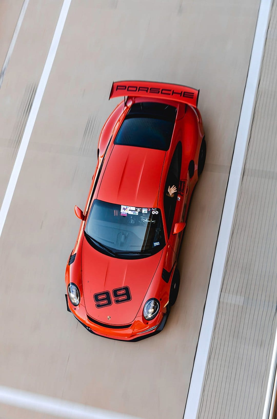 Red Porsche 99 racing car with a rear wing sits on a light gray track during a high-speed automotive event.