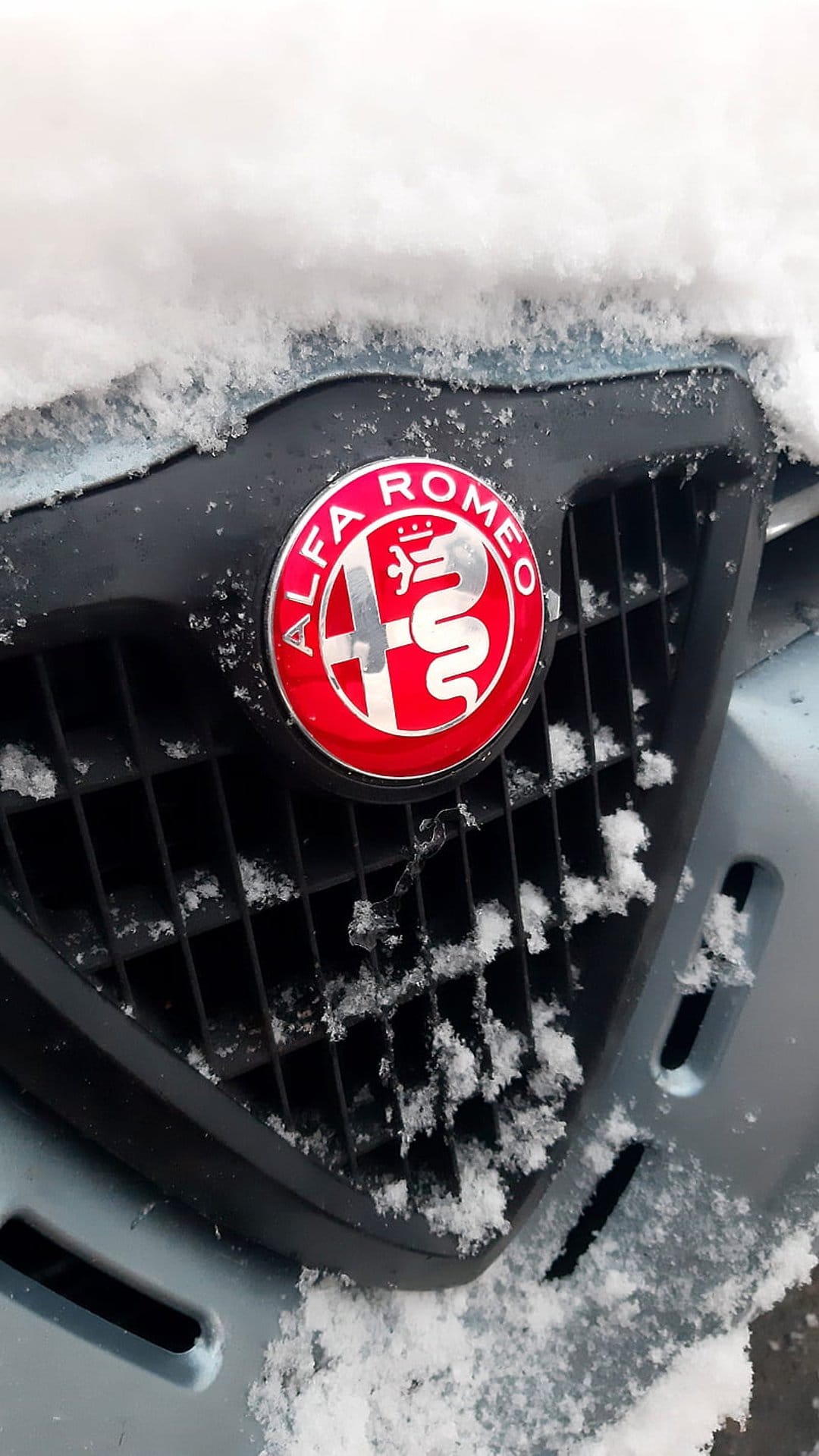 Alfa Romeo emblem centered on a black mesh car grille covered in crystalline snow and frosty ice particles.