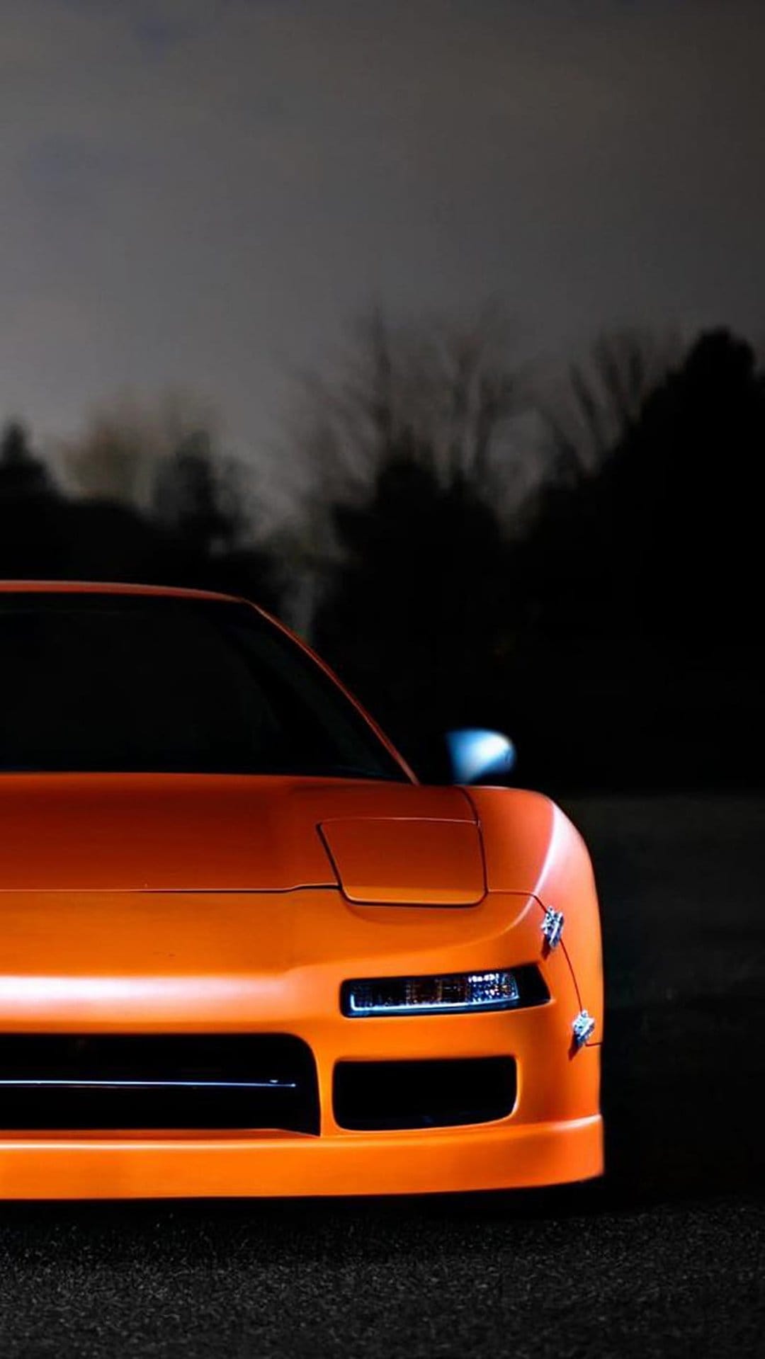 Orange Honda NSX sports car with glowing blue headlights parked on dark asphalt with silhouetted trees.