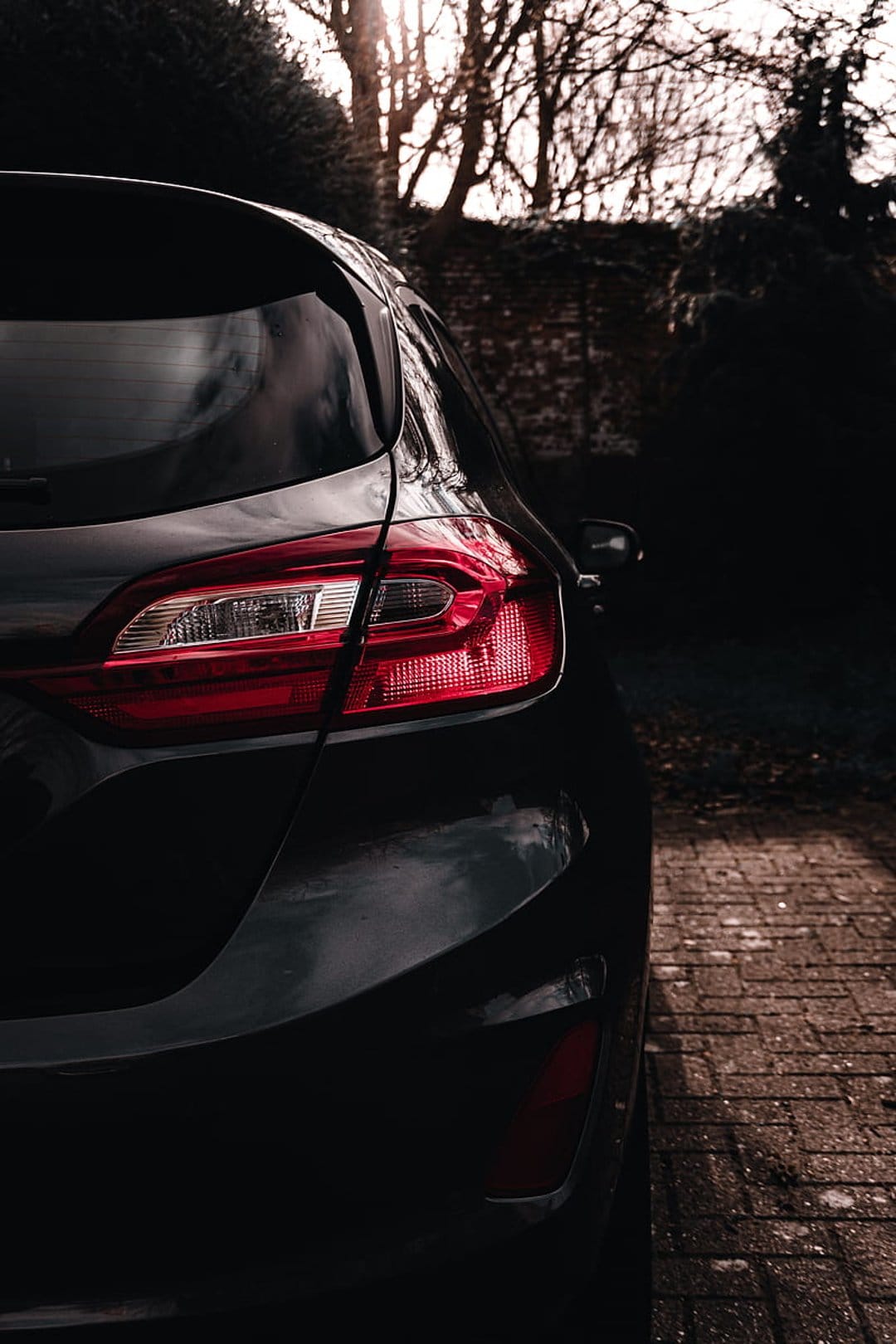 Red car taillight and sleek dark bodywork on brick pavement with a blurred background of trees and architecture.