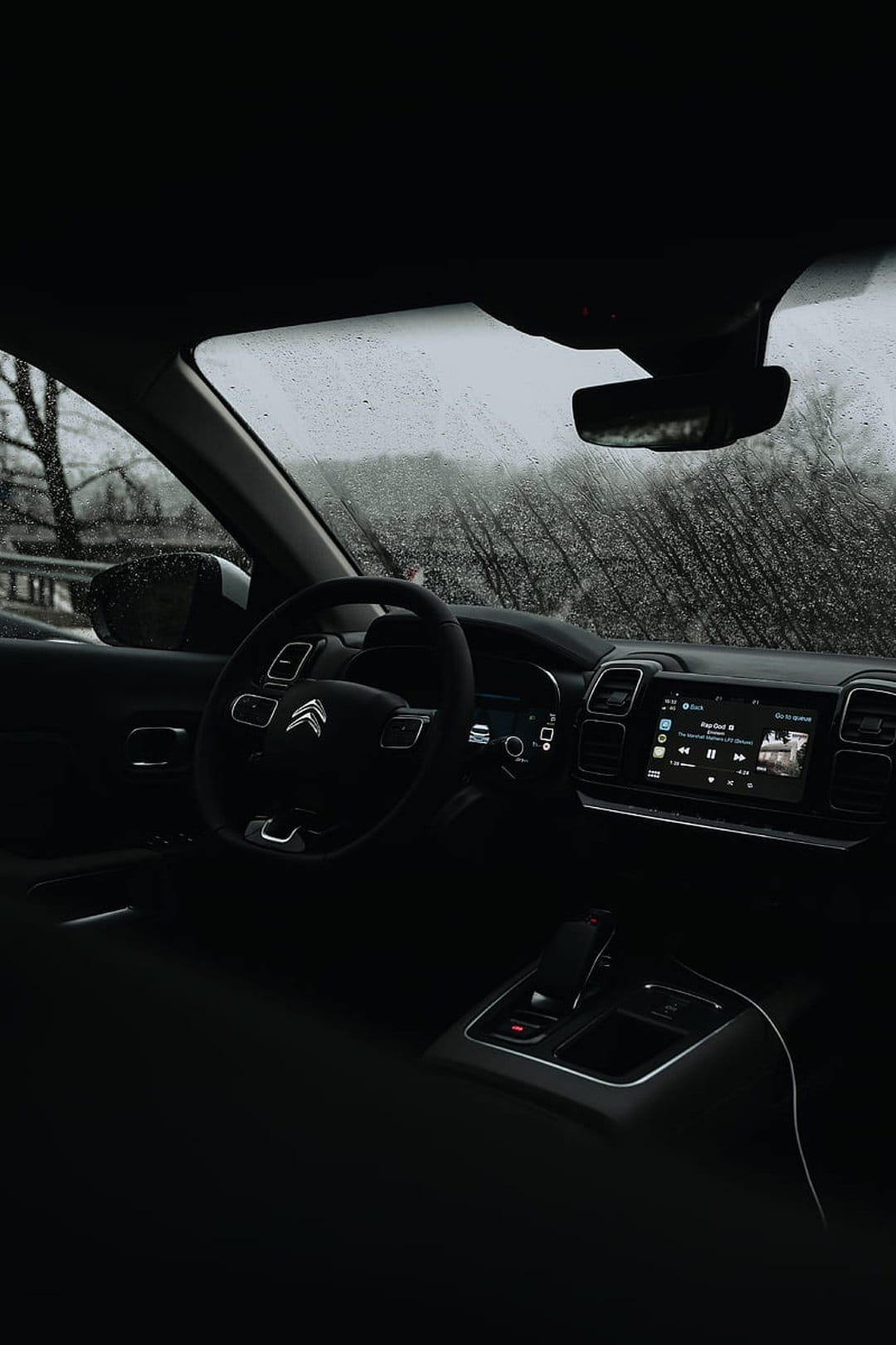 Raindrops on a car windshield frame a dark dashboard and glowing screen with blurred trees in the distance.