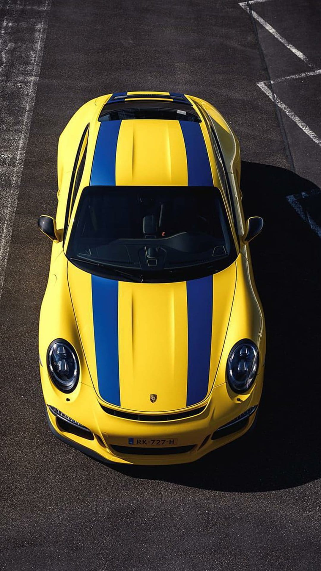 Yellow Porsche 911 sports car with blue racing stripes parked on dark asphalt viewed from a top-down perspective.