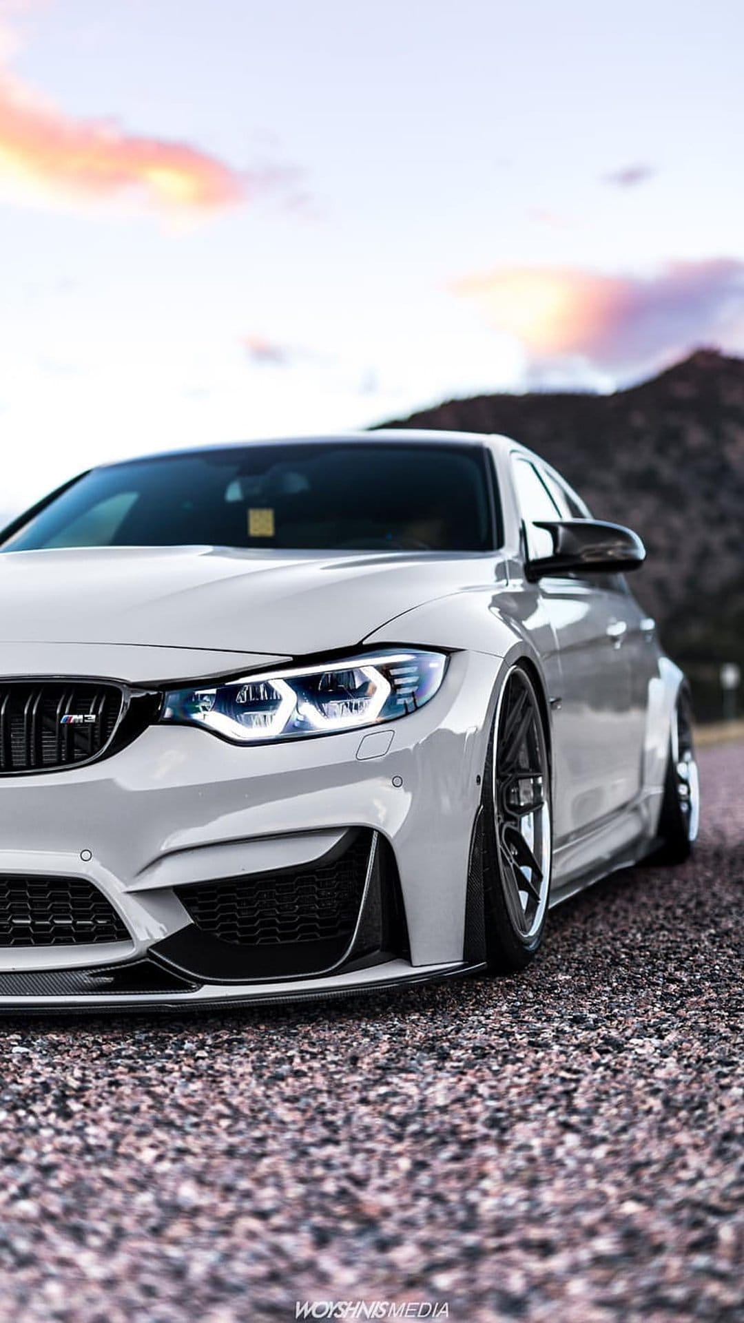White BMW M4 widebody with glowing headlights parked on a gravel road during a vibrant orange and purple sunset.