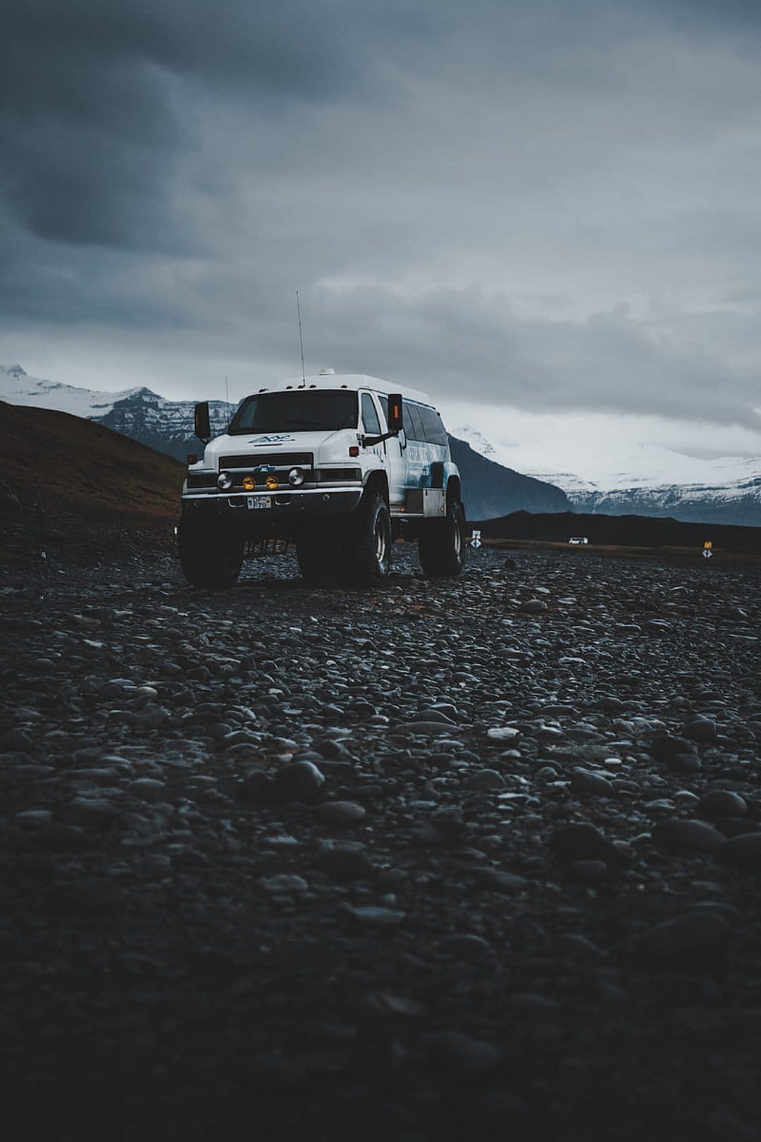 White expedition van with oversized tires parked on dark volcanic rocks before snowy mountains under storm clouds.