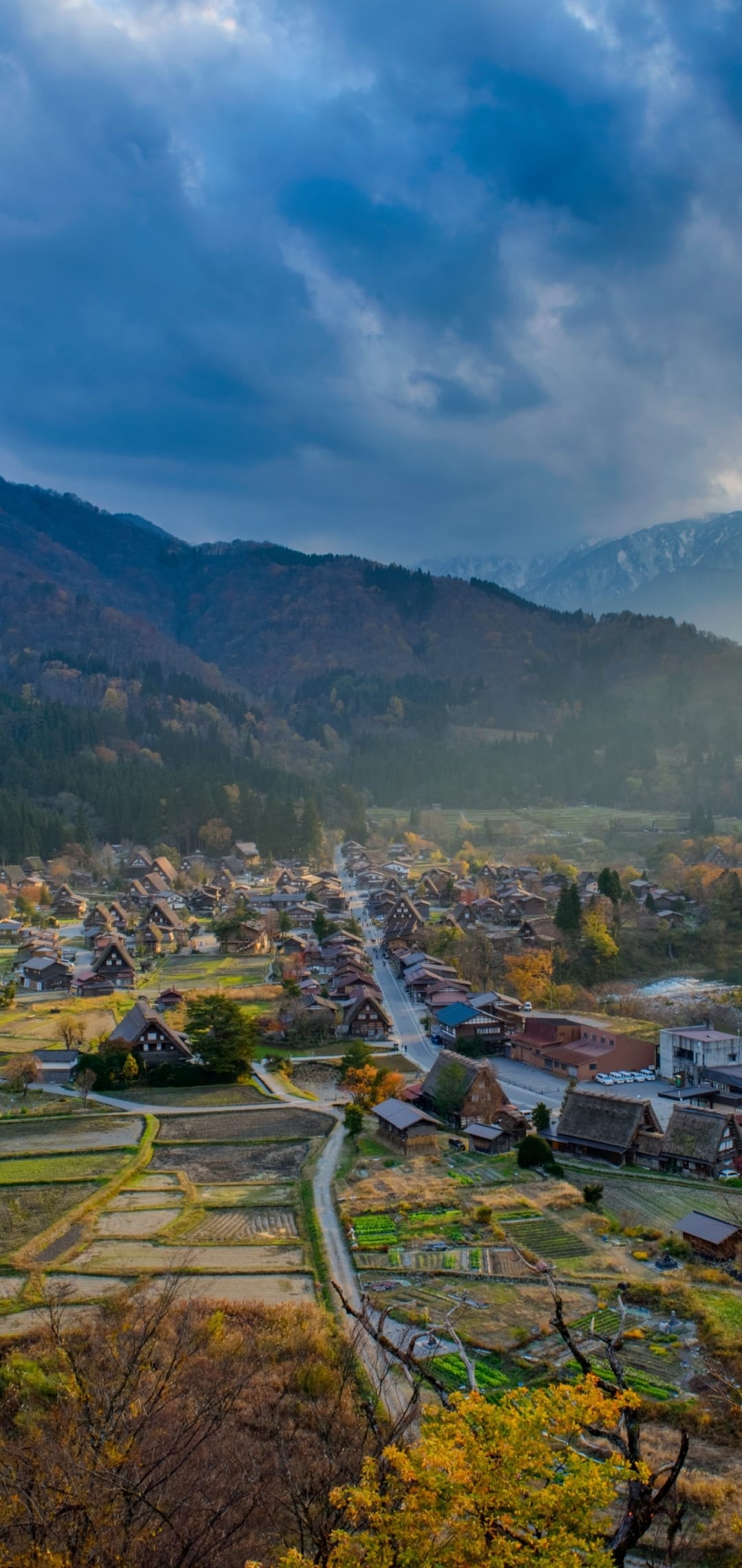 Thatched roof houses nestle in a mountain valley surrounded by orange autumn trees and golden harvest fields.