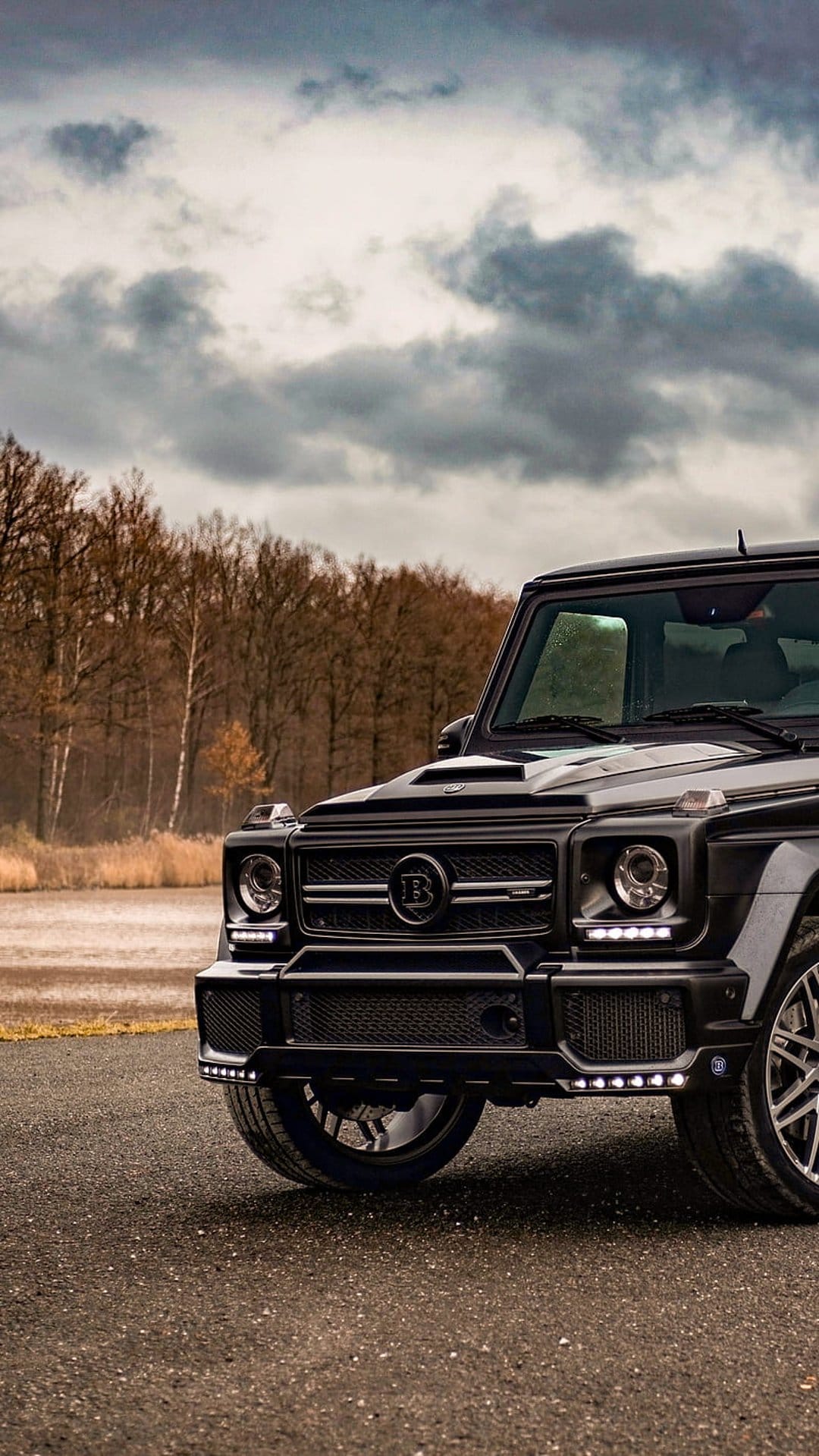 Black Brabus G-Wagon SUV parked on a road beside a lake under a cloudy sky in a low-angle vertical shot.