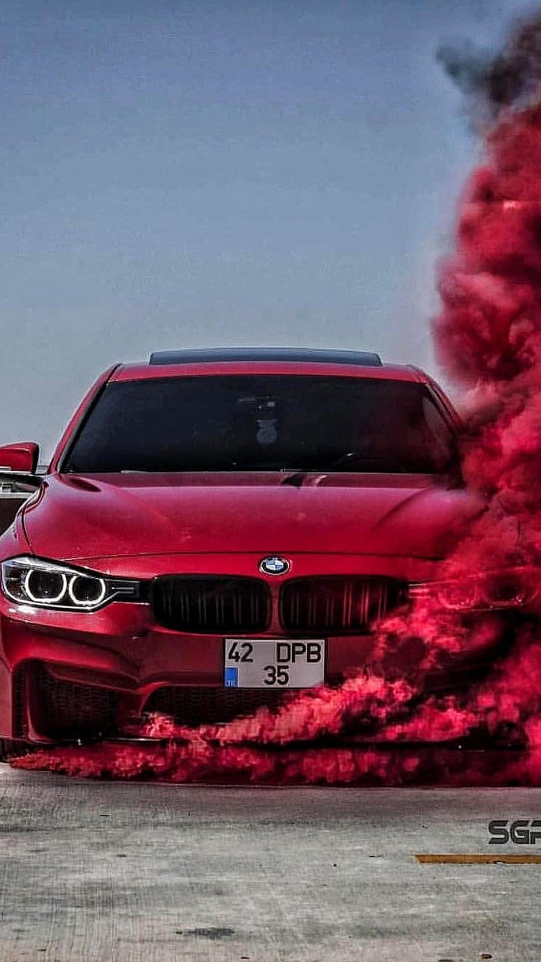 Red BMW sedan surrounded by thick plumes of crimson smoke on a sunny urban street under a clear blue sky.