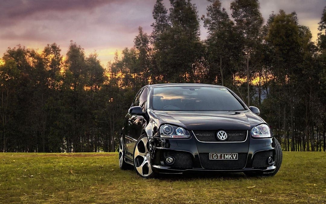 Black Volkswagen GTI parked on a grassy field with a purple and orange sunset sky behind tall forest trees.