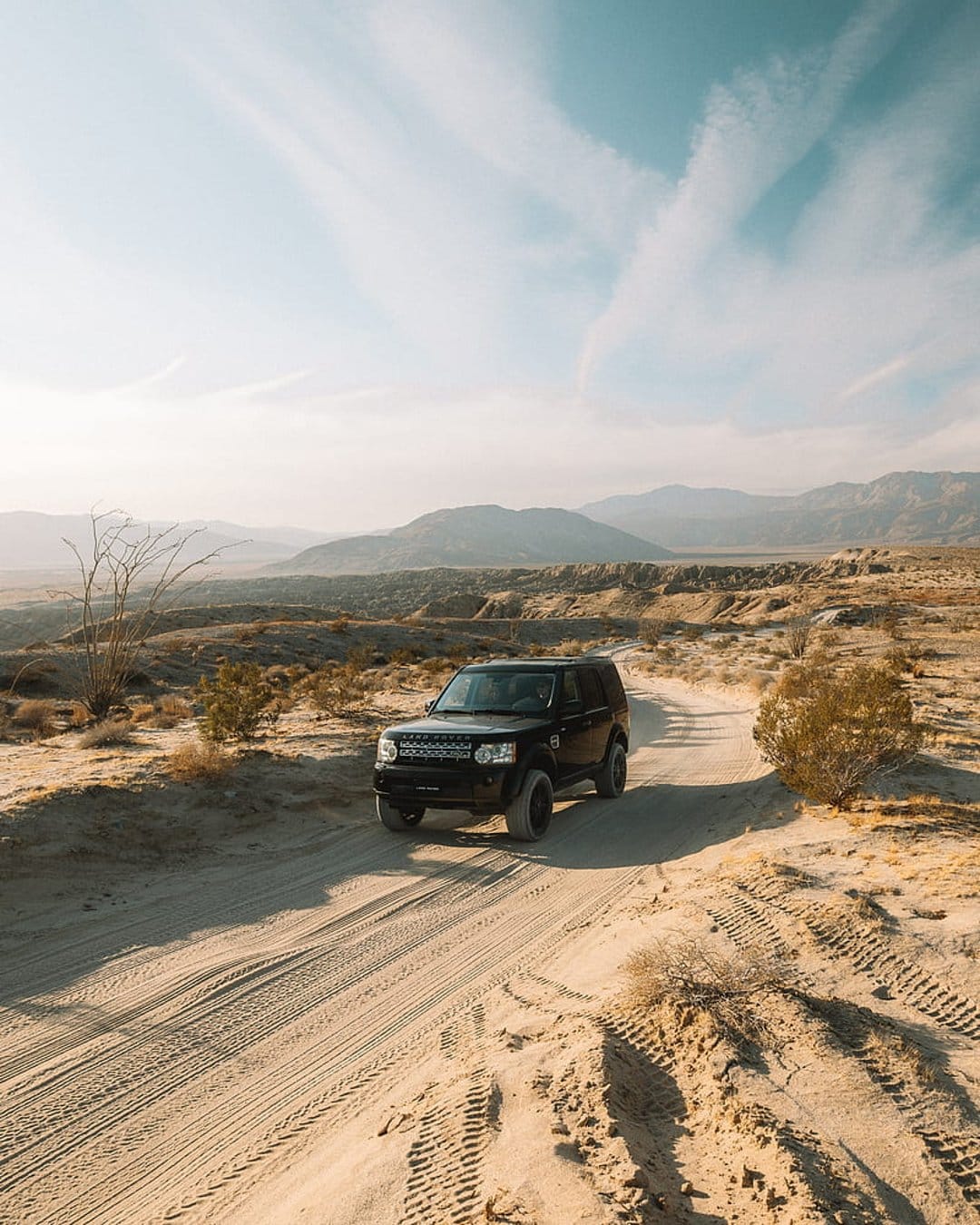 Black SUV driving on a winding sandy track through a vast desert with distant mountains under a cloudy sky.