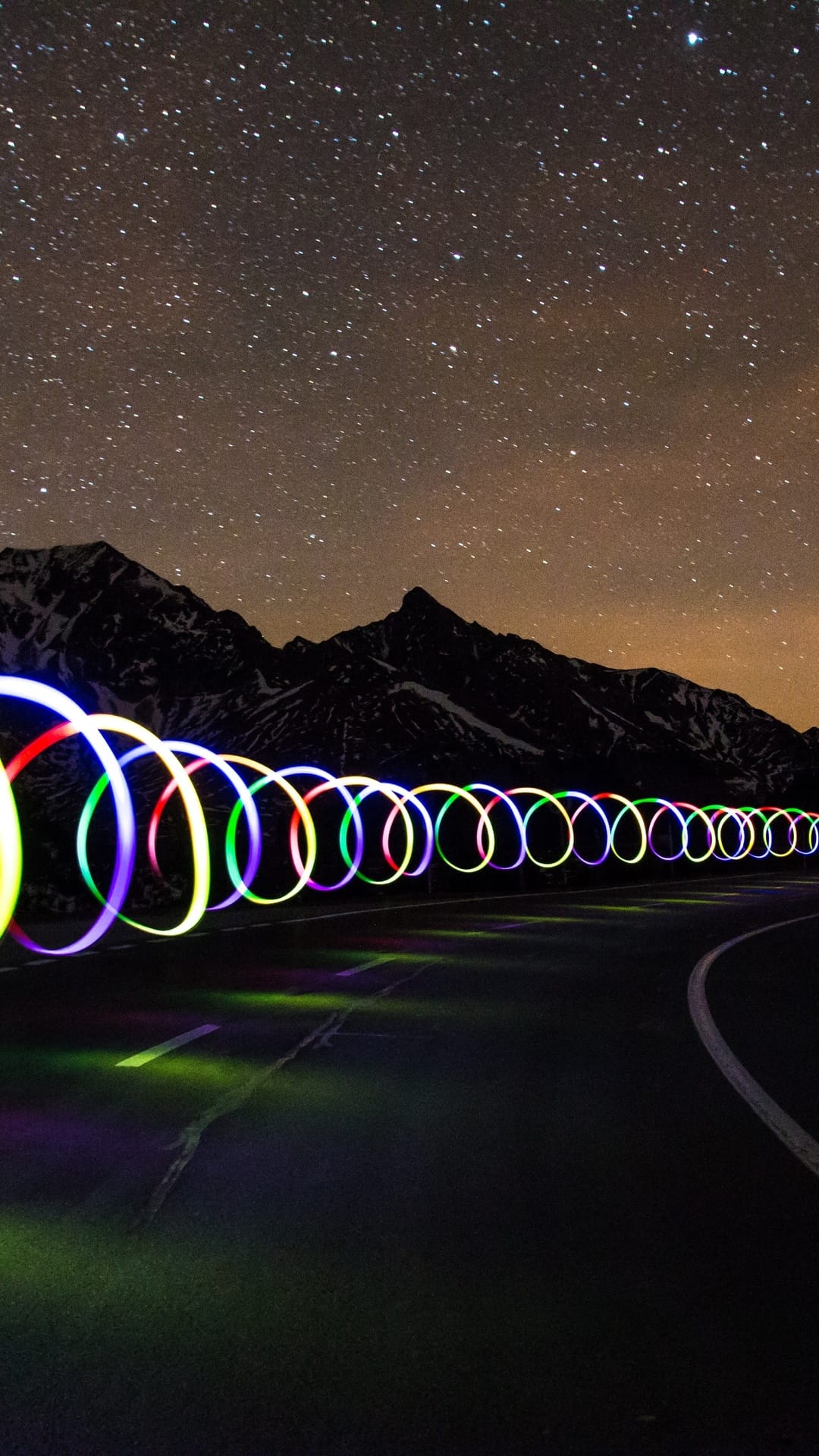 Multicolored neon circular light trails spiral along a mountain road beneath a star-filled sky and jagged peaks.