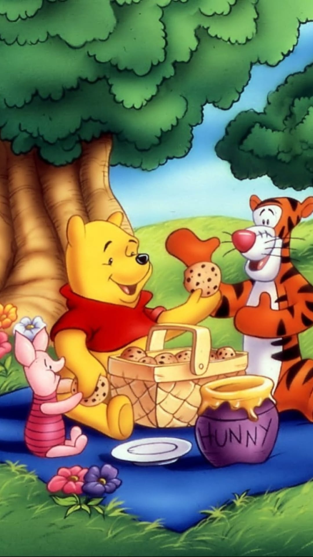 Winnie the Pooh, Tigger, and Piglet sit on a picnic blanket eating cookies under a large leafy tree.