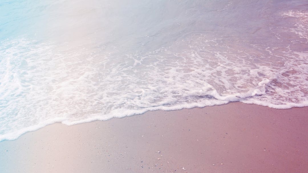 Pastel sea foam glides across a pale sandy beach during a soft sunset, creating a tranquil coastal scene.