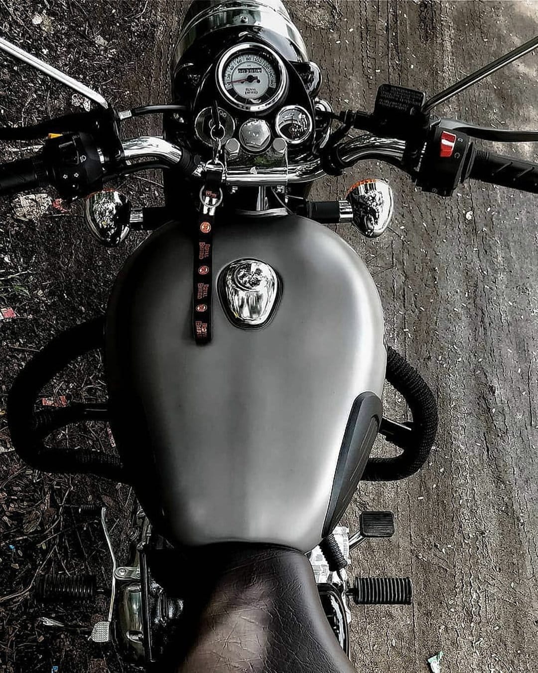 Grey motorcycle fuel tank and chrome handlebars featuring a round analog speedometer and leather keychain.