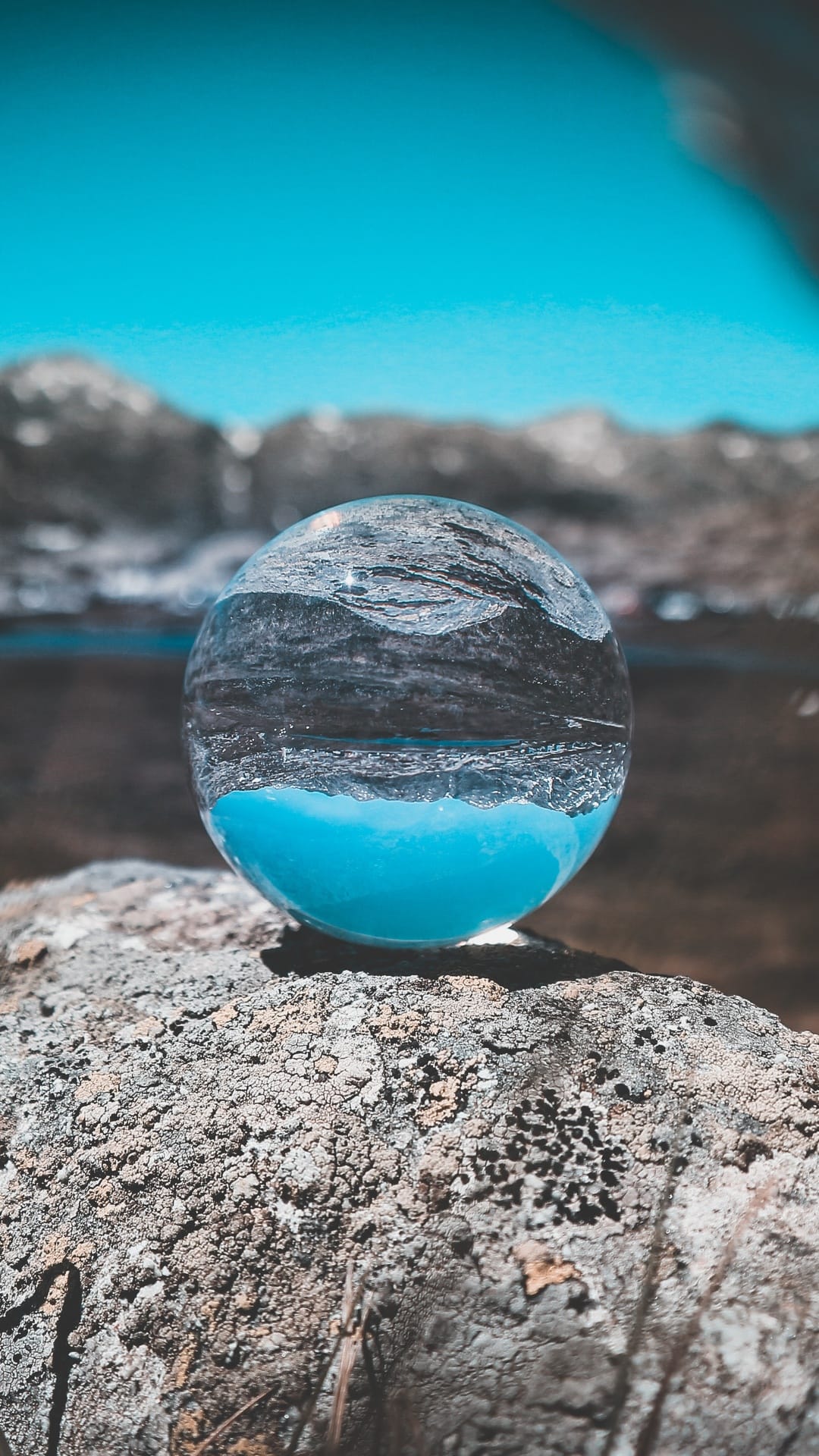 Crystal sphere on a rock captures an inverted reflection of blue mountains and a turquoise lake under a clear sky.