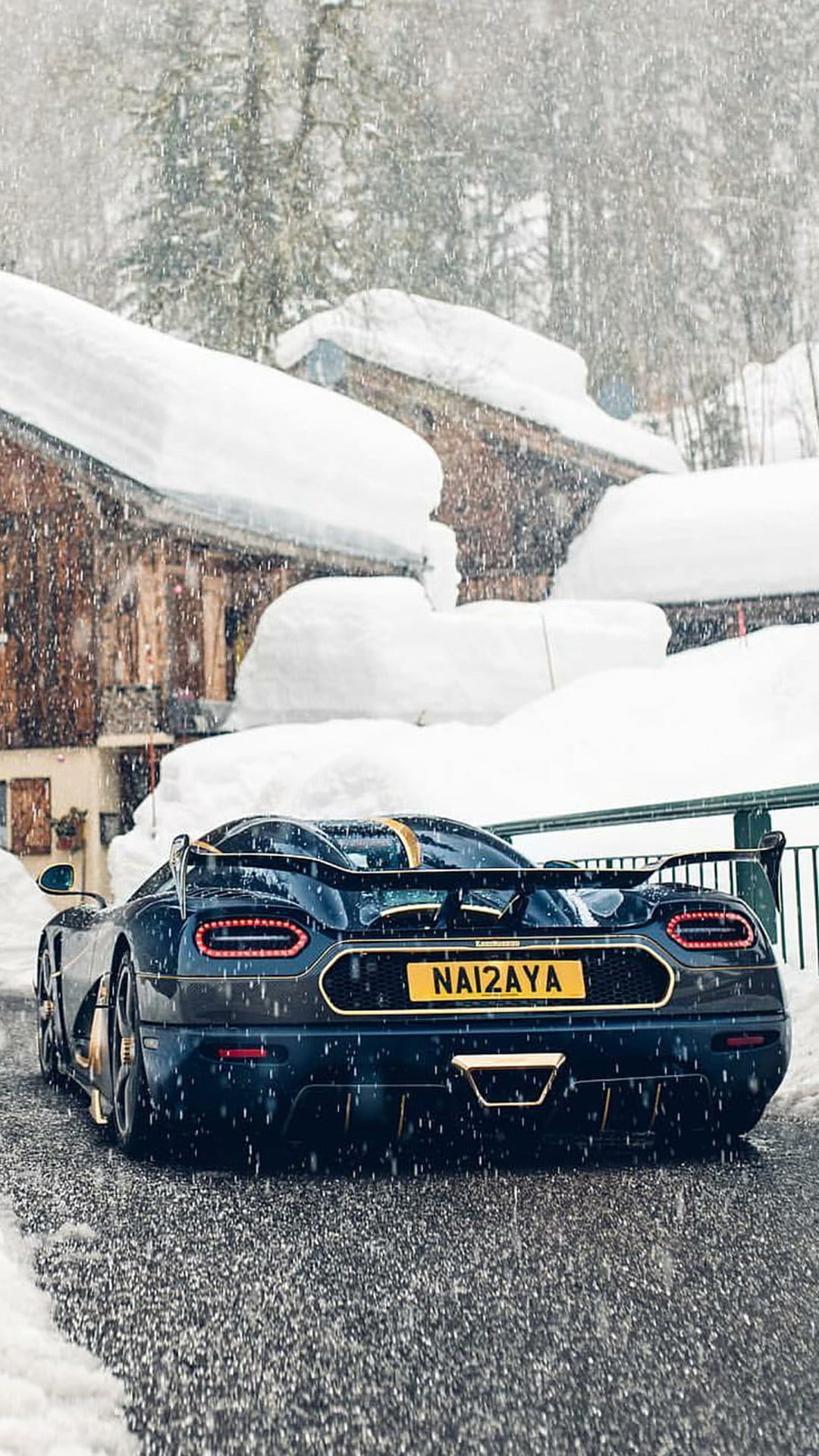 Dark blue Koenigsegg with gold trim on a wet snowy road near frost-covered wooden chalets during a light snowfall.