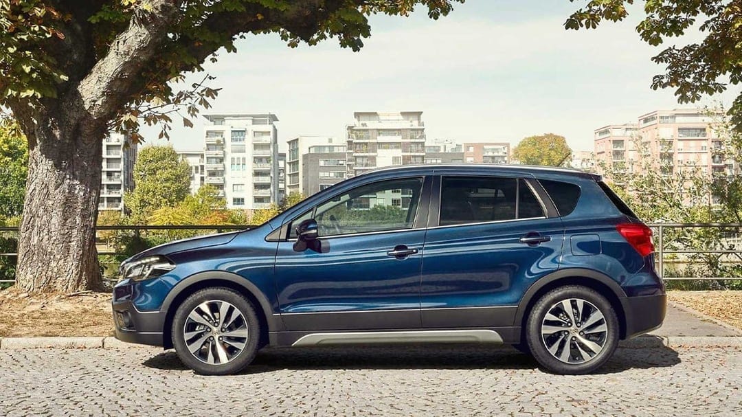Blue Suzuki S-Cross SUV parked in profile on a cobblestone street beneath a large green tree in an urban setting.