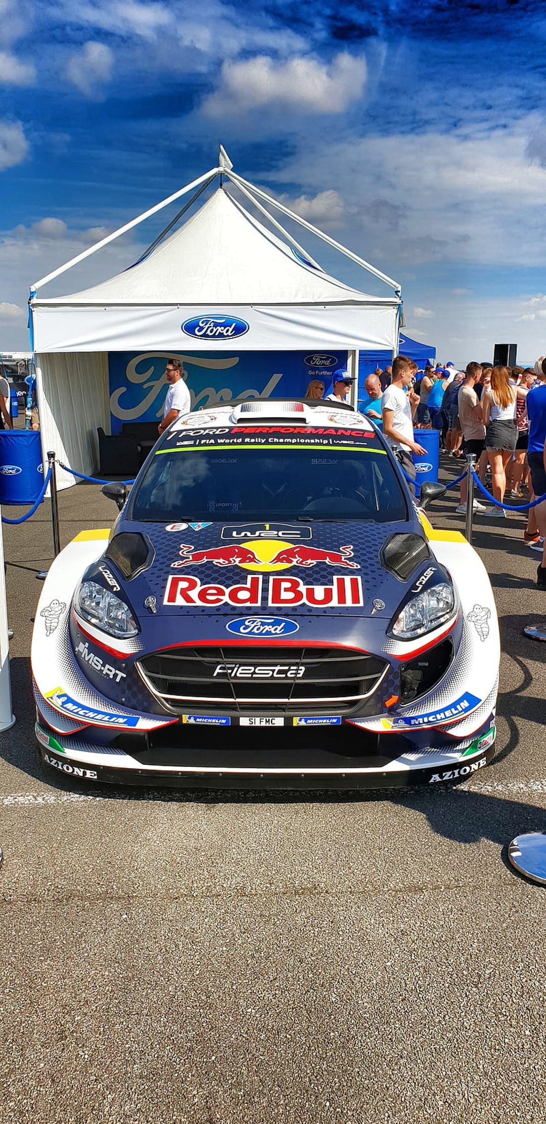Ford Fiesta WRC rally car featuring Red Bull decals and Michelin tires under an exhibition tent outdoors.