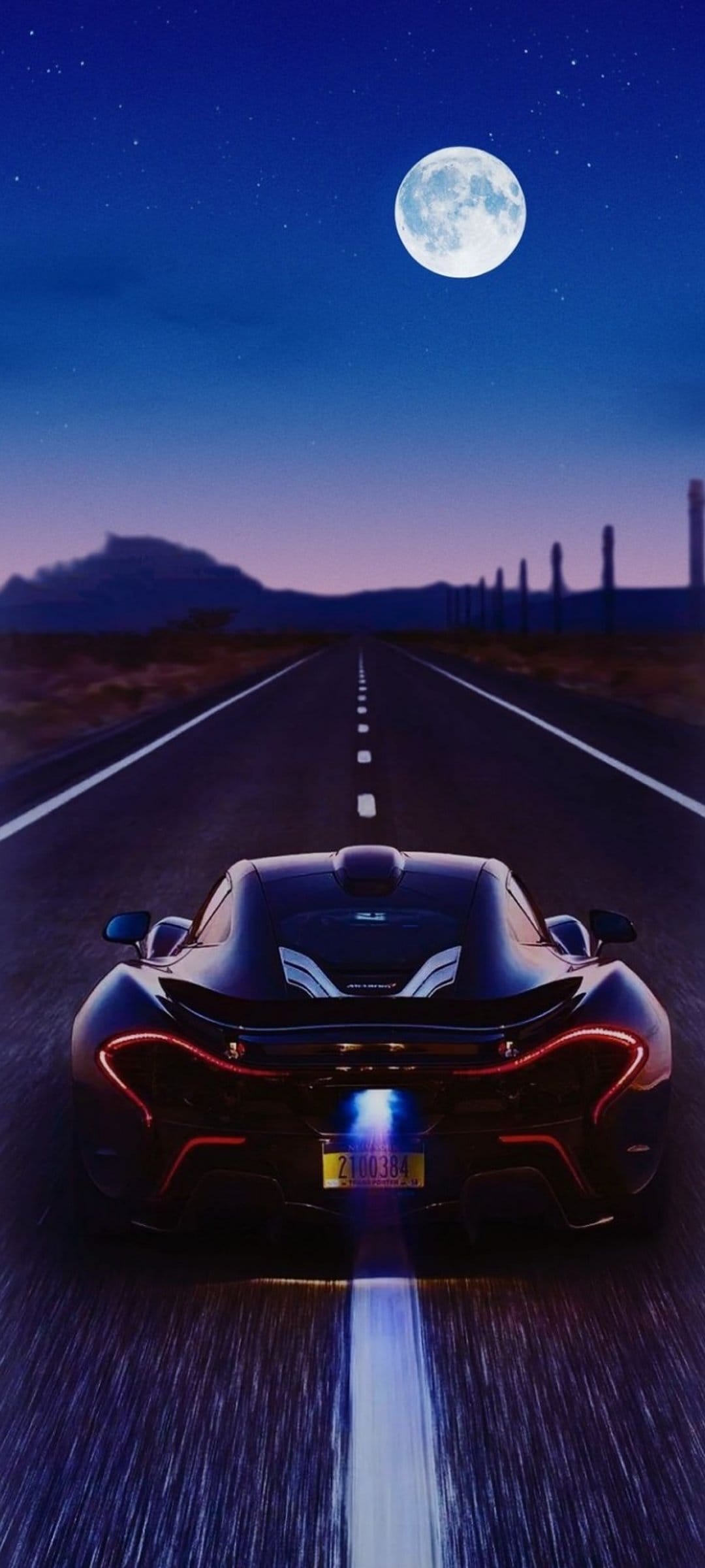 Black supercar with red taillights speeds along a desert highway under a bright full moon and starry night sky.