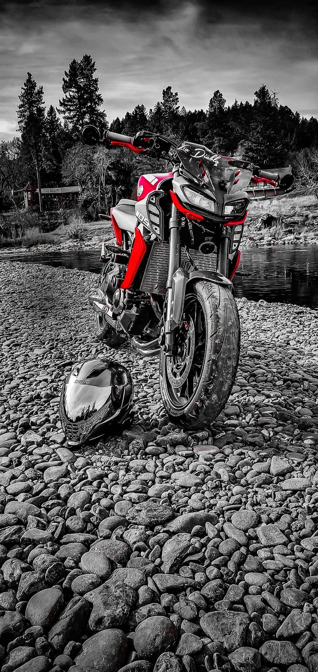 Red sport motorcycle and matching helmet rest on a grey rocky riverbed with a desaturated forest background.