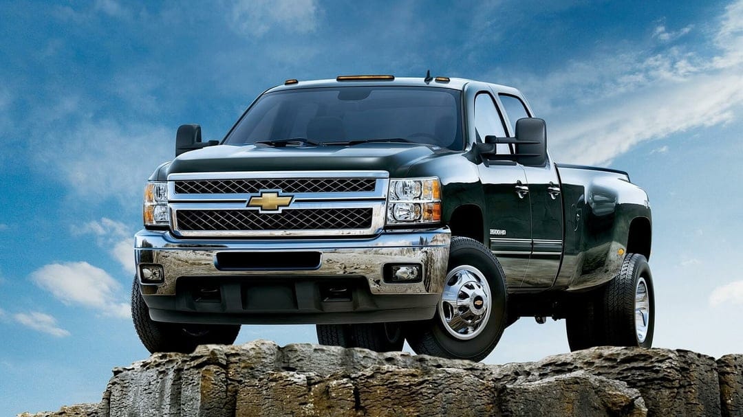 Dark Chevrolet Silverado dually truck on a rocky cliff under a blue sky with chrome grille and headlights.