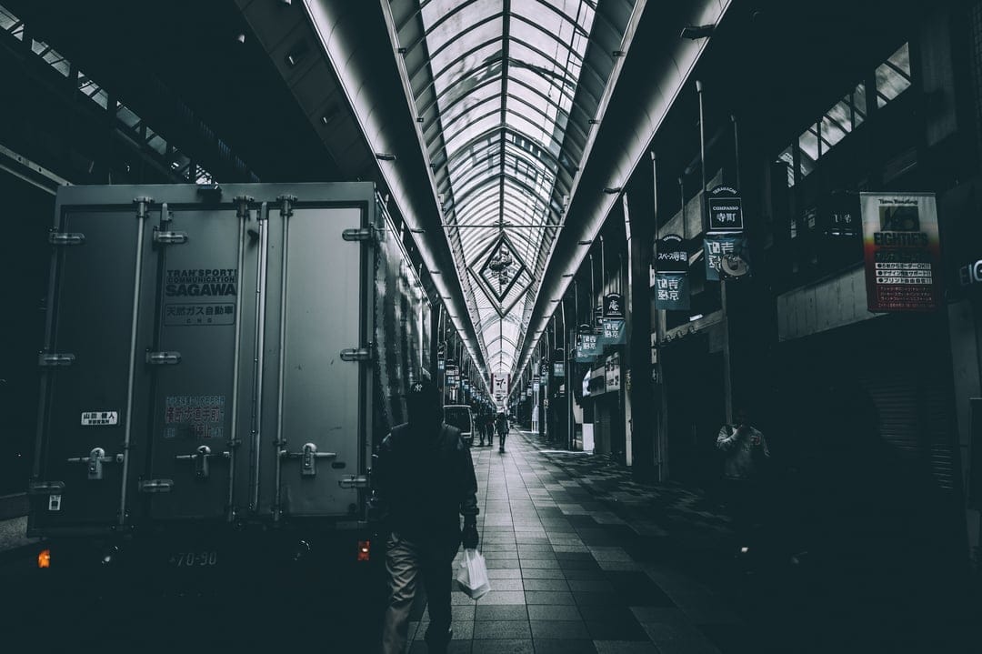 Lone man walks through a dark arcade with a geometric glass roof and dramatic noir-style urban shadows.