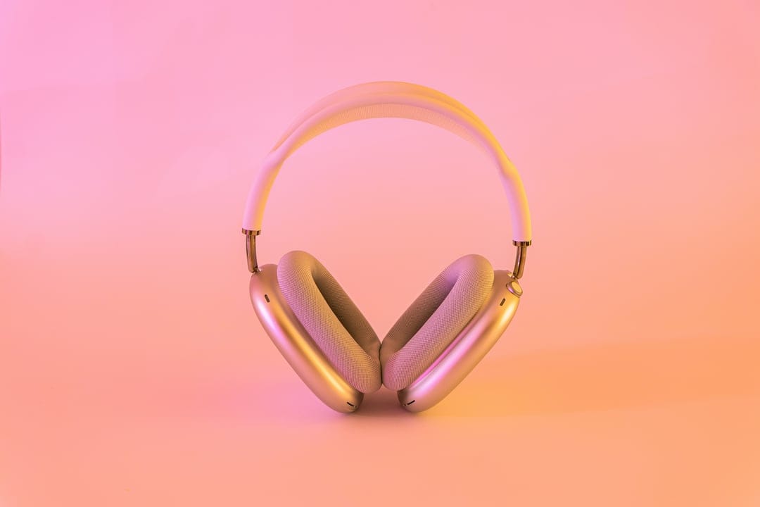 Silver over-ear headphones centered on a soft pink and orange gradient background with subtle metallic highlights.