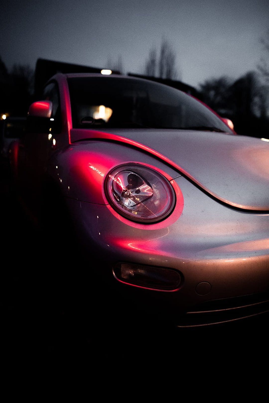 Silver Volkswagen Beetle with red neon reflections on its fender and headlight against a dark background.