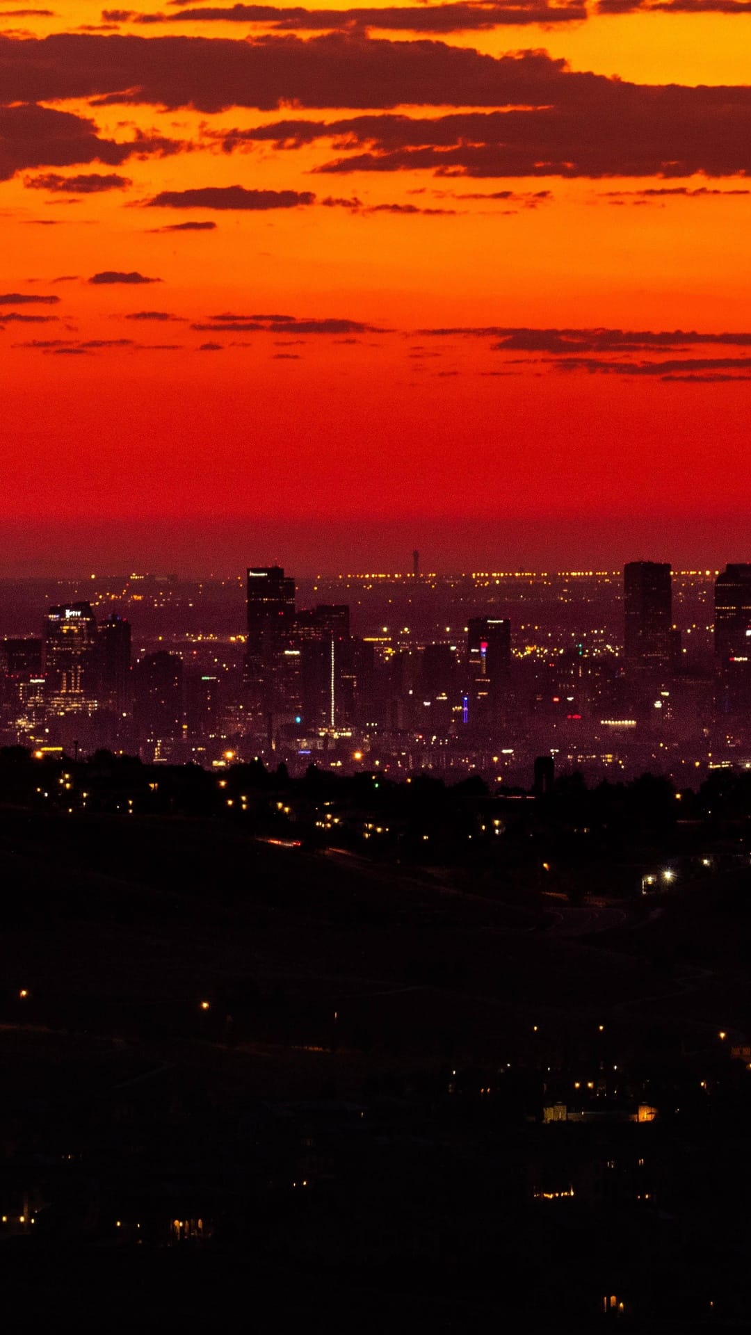 City skyline silhouettes against a fiery red and orange sunset sky with glowing urban lights and skyscrapers.