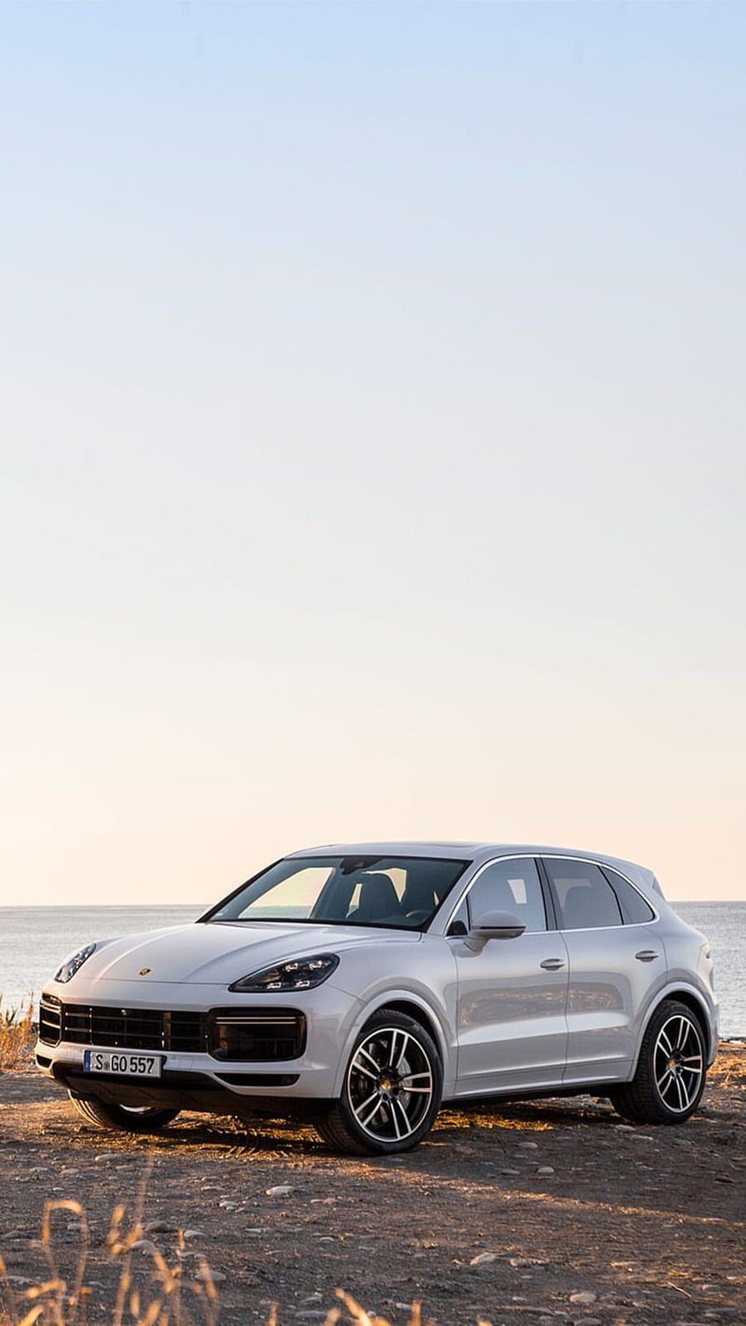 Silver Porsche Cayenne SUV parked on a gravel shore against a hazy ocean sunset in a vertical composition.