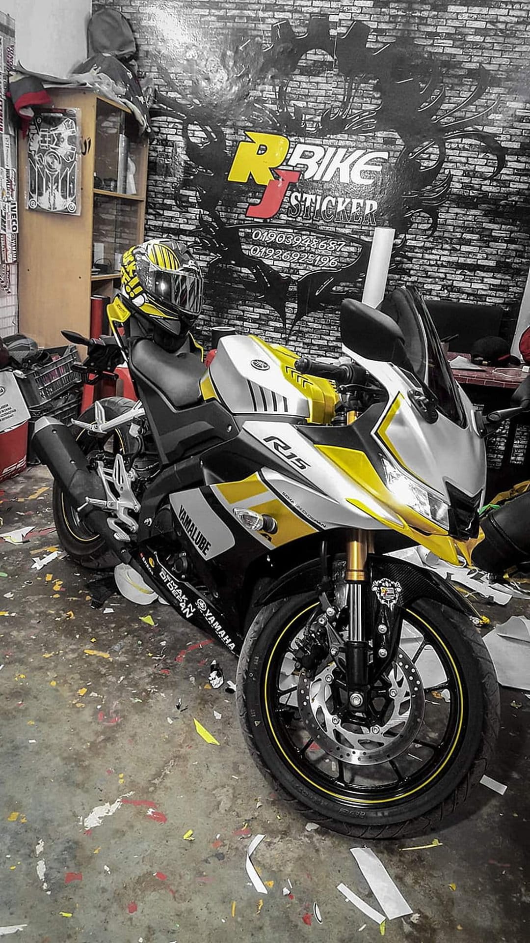 Yamaha R15 motorcycle with yellow and silver decals parked in a workshop against a brick wall background.
