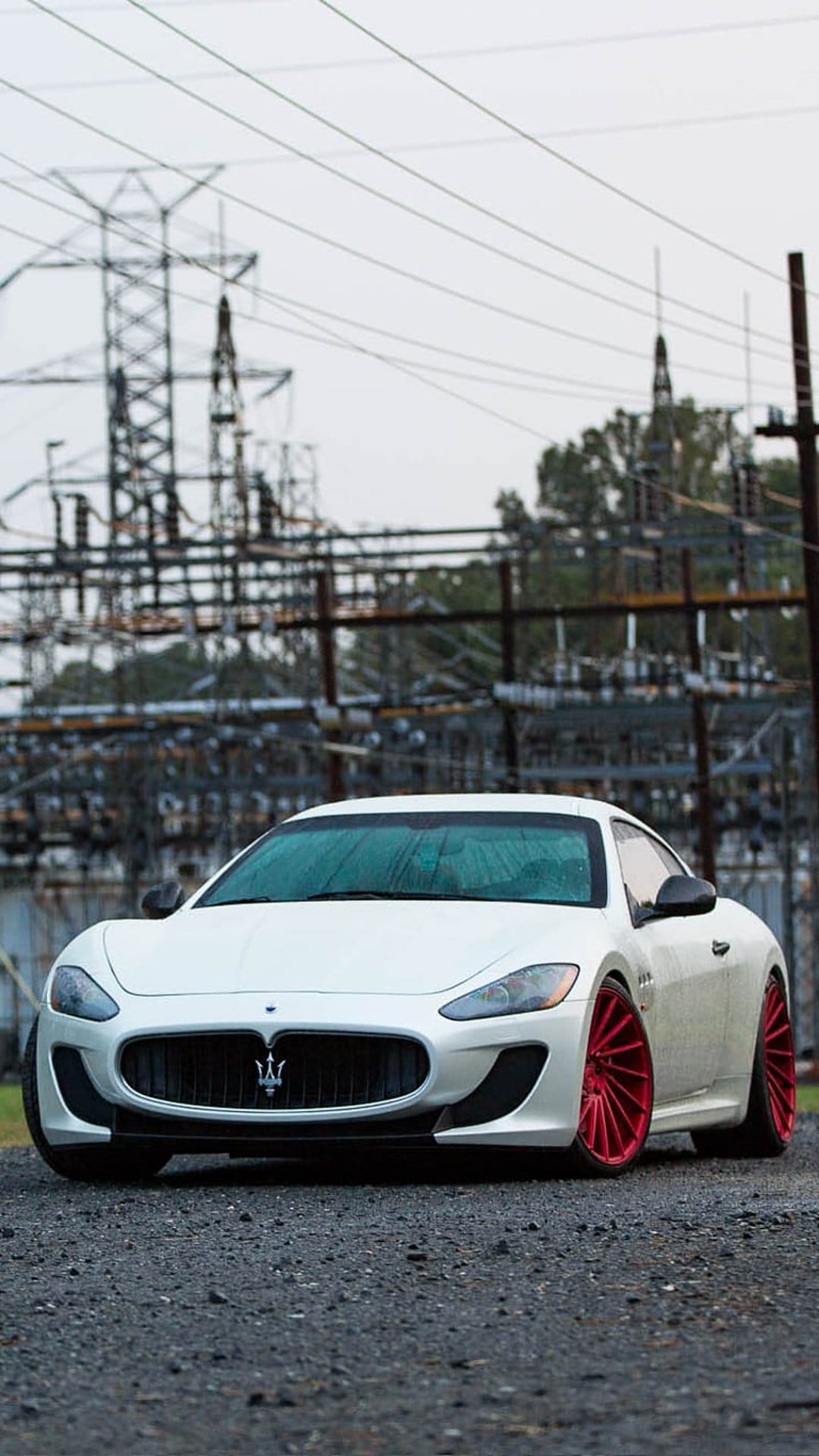 White Maserati GranTurismo with red wheels parked on gravel beneath industrial power towers and an overcast sky.