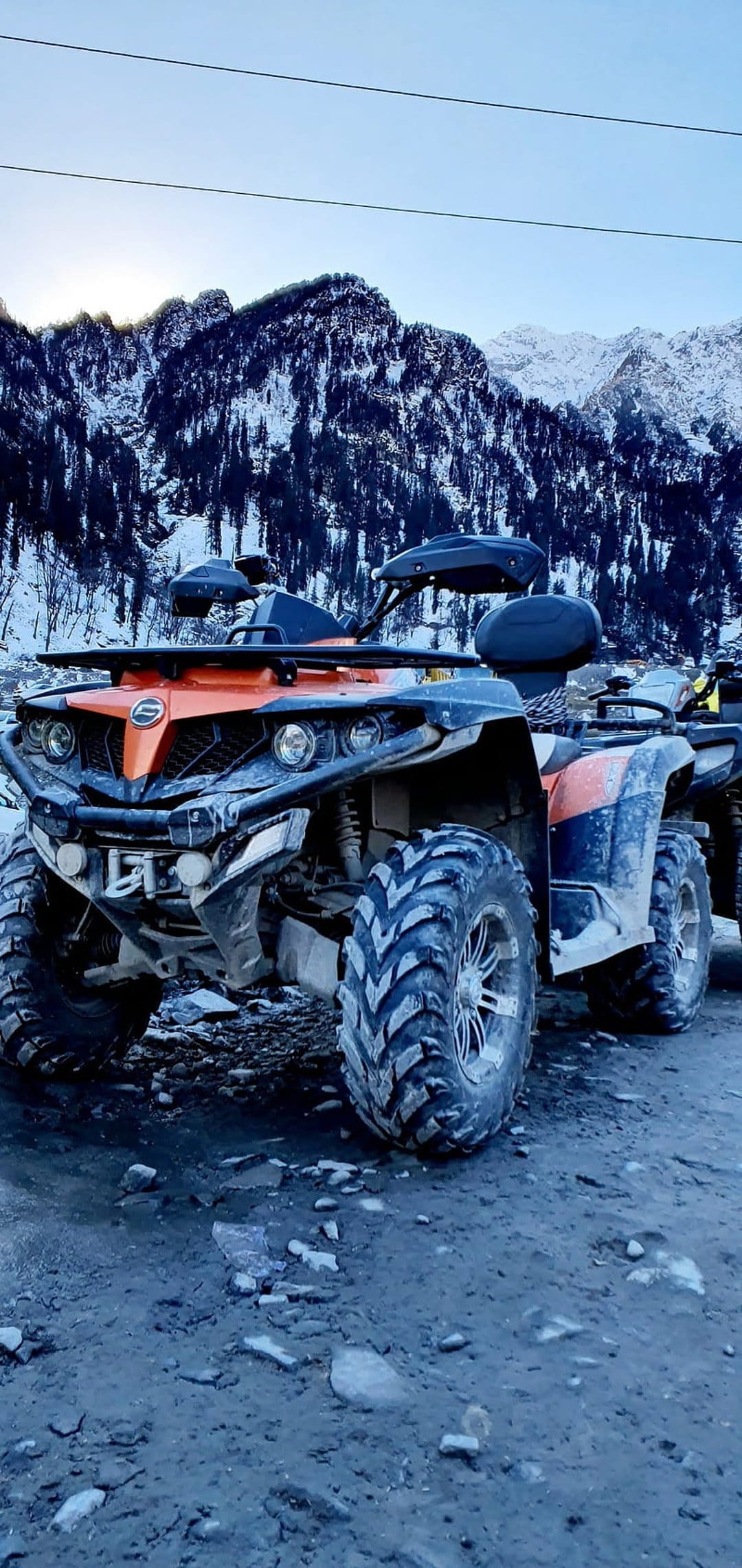 Orange and black ATV on a rocky ridge with snow-covered mountain peaks under a clear blue alpine winter sky.