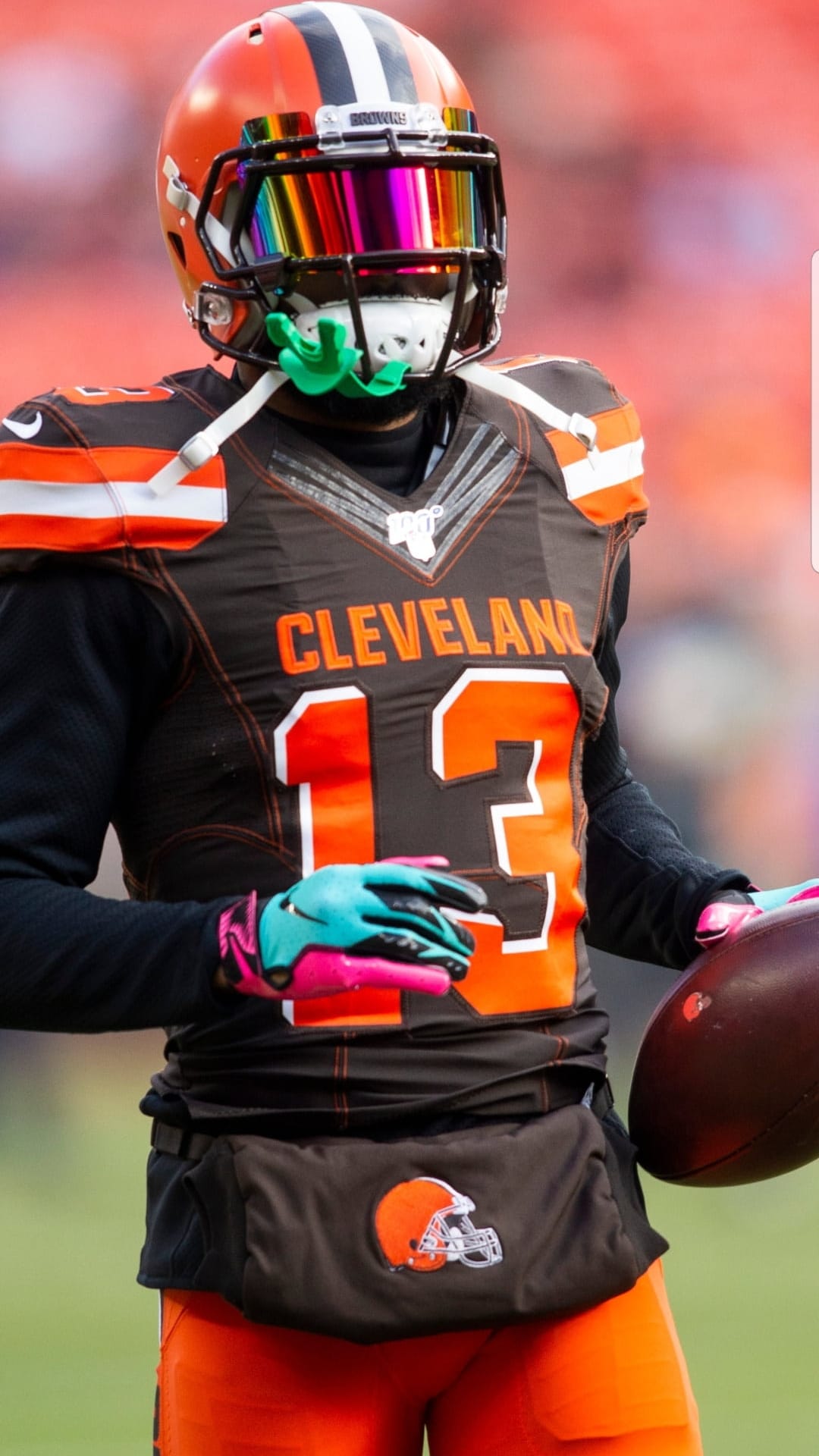 Cleveland Browns football player wearing a multicolored helmet visor and turquoise gloves holding a football.