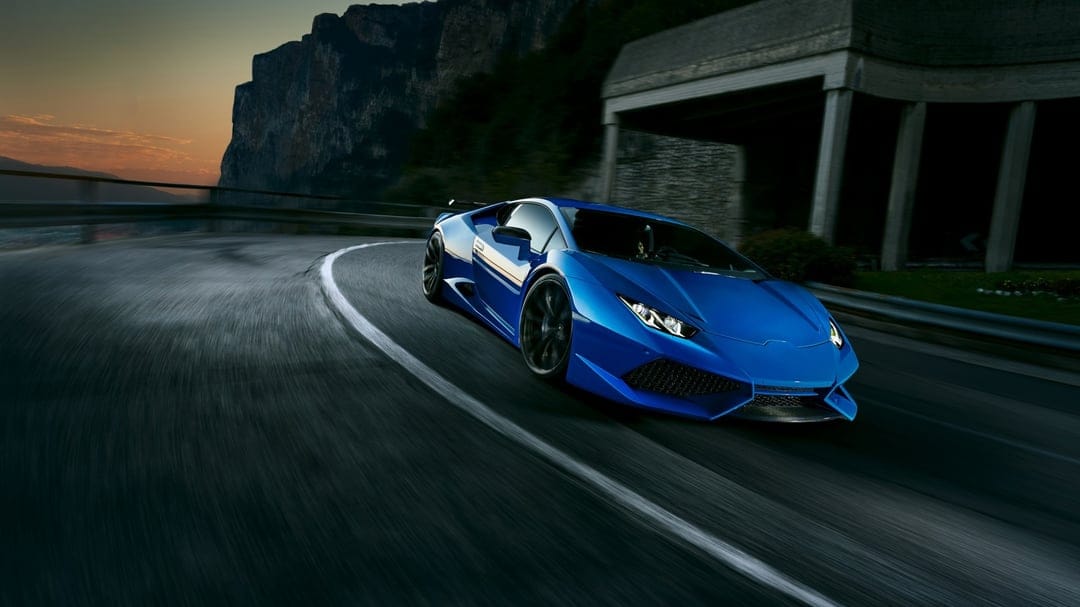 Blue supercar speeding through a curved mountain road at sunset with heavy motion blur and rugged cliffs.
