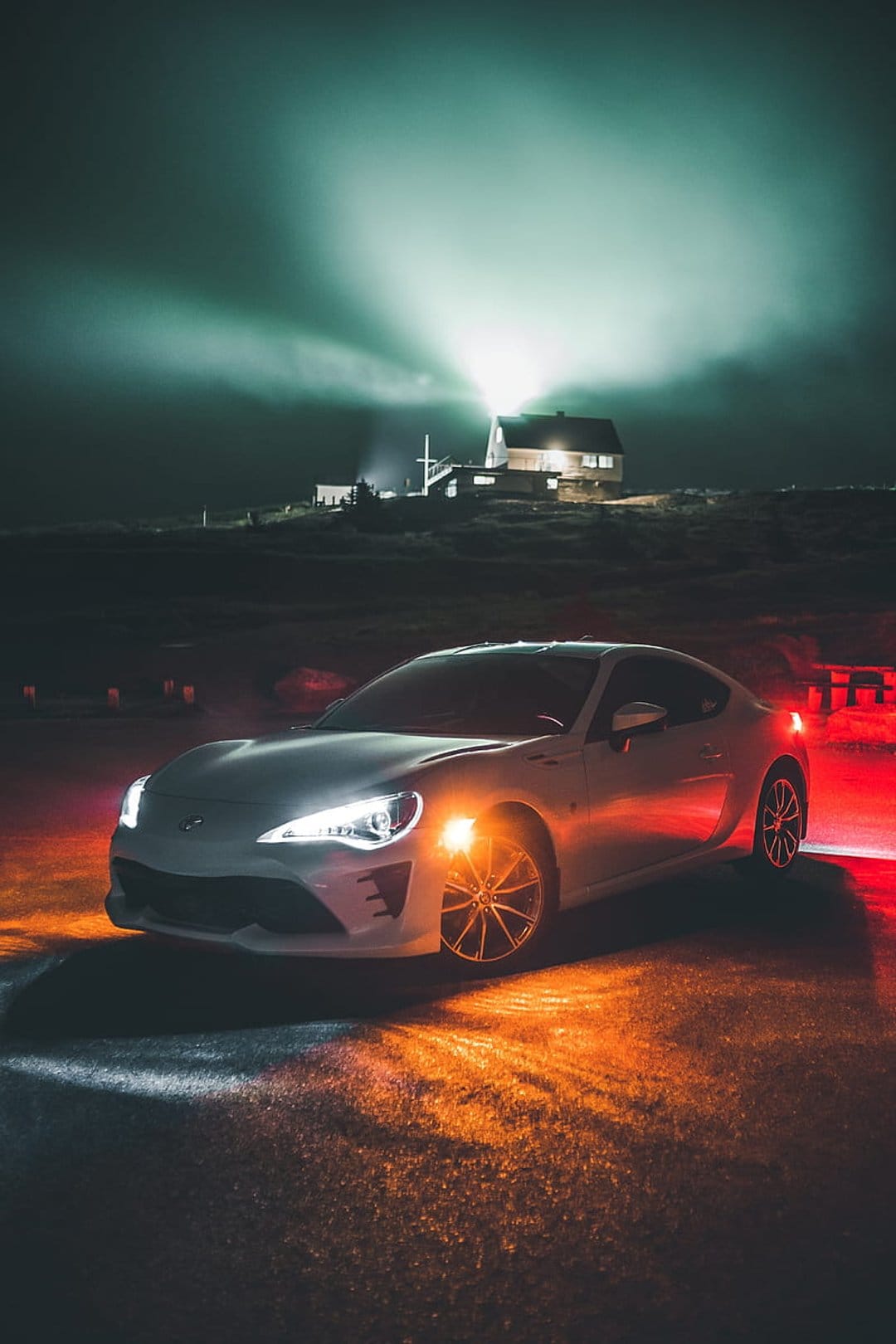 White sports car with glowing headlights parked at night as a lighthouse beam cuts through the misty blue sky.